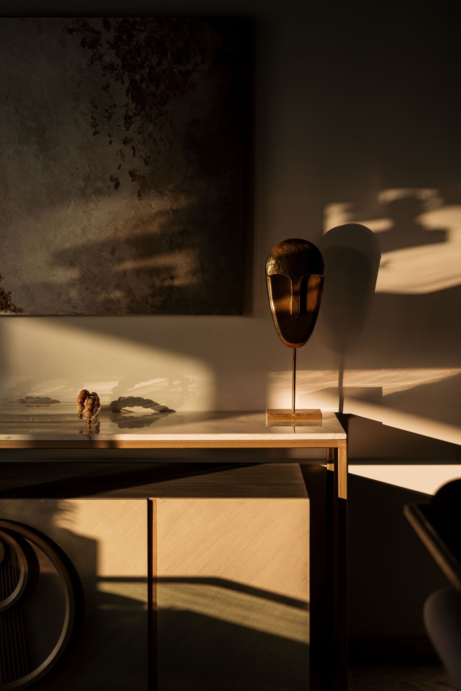 Dark dramatic vignette featuring a bronze African mask sculpture on a gold console table beneath a moody abstract painting.