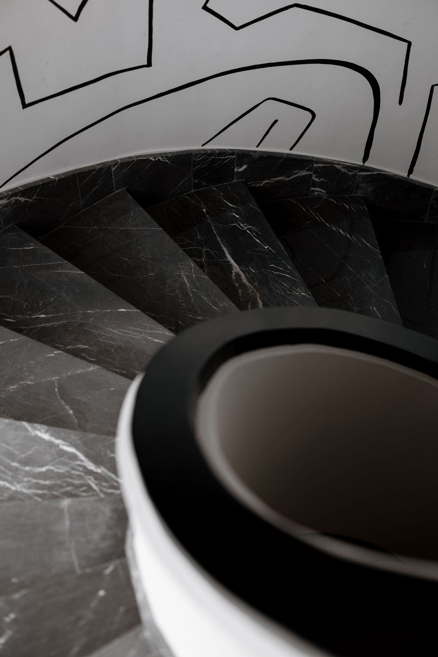 Aerial view of dark marble spiral staircase with abstract black line mural on white walls.
