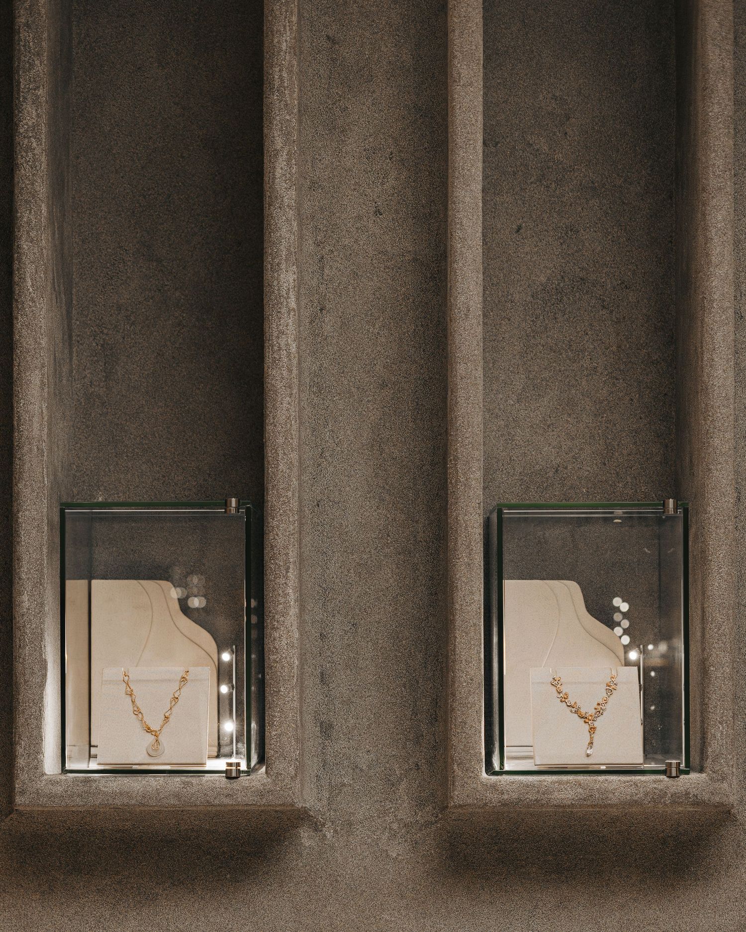 Two illuminated wall niches in textured concrete displaying elegant gold necklaces in glass cases at a jewelry store.