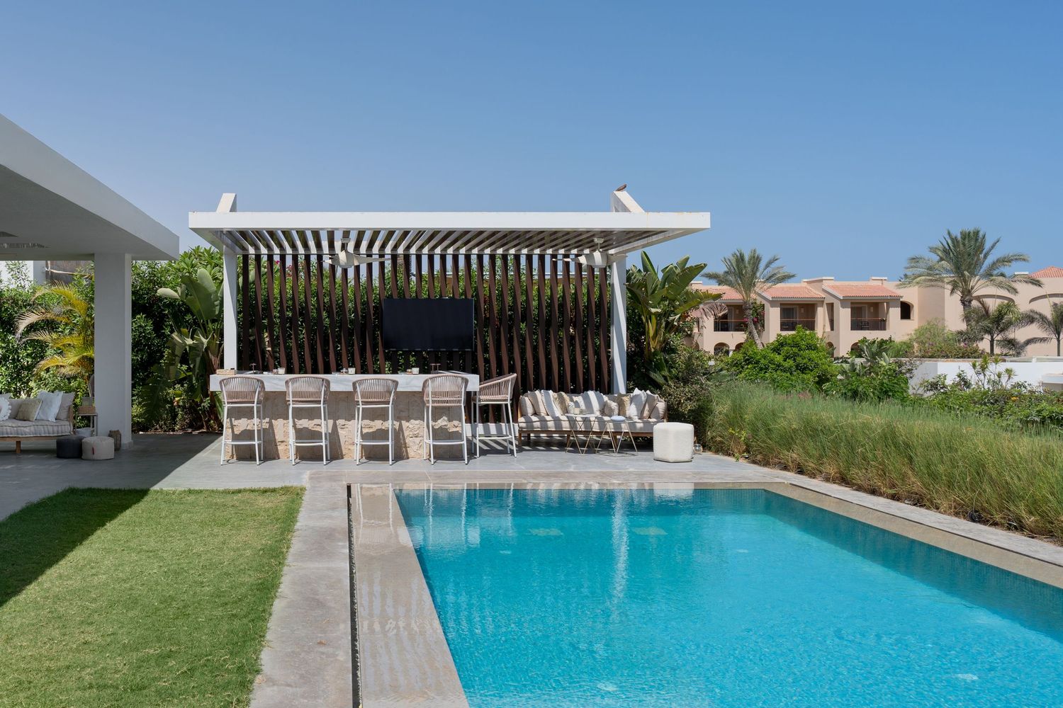 Sleek outdoor pool area with white pergola, lounge chairs, manicured lawn, and palm trees under blue sky.