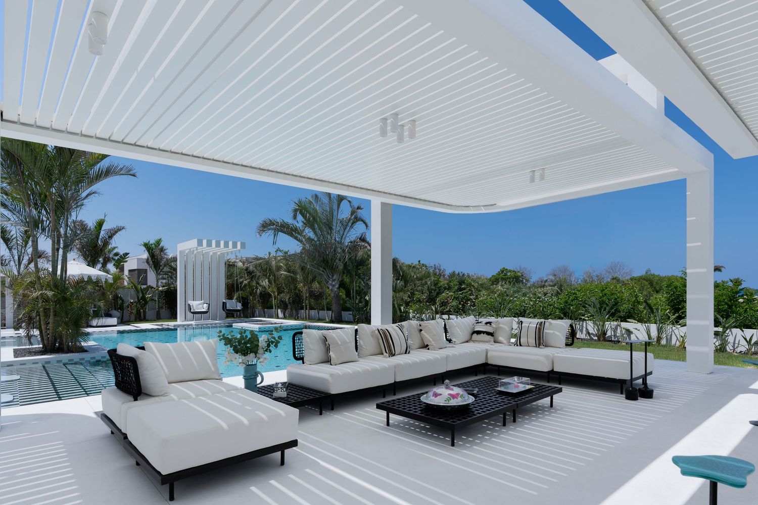Spacious white outdoor living area with sectional sofa, black coffee table, pool view, and pergola ceiling.