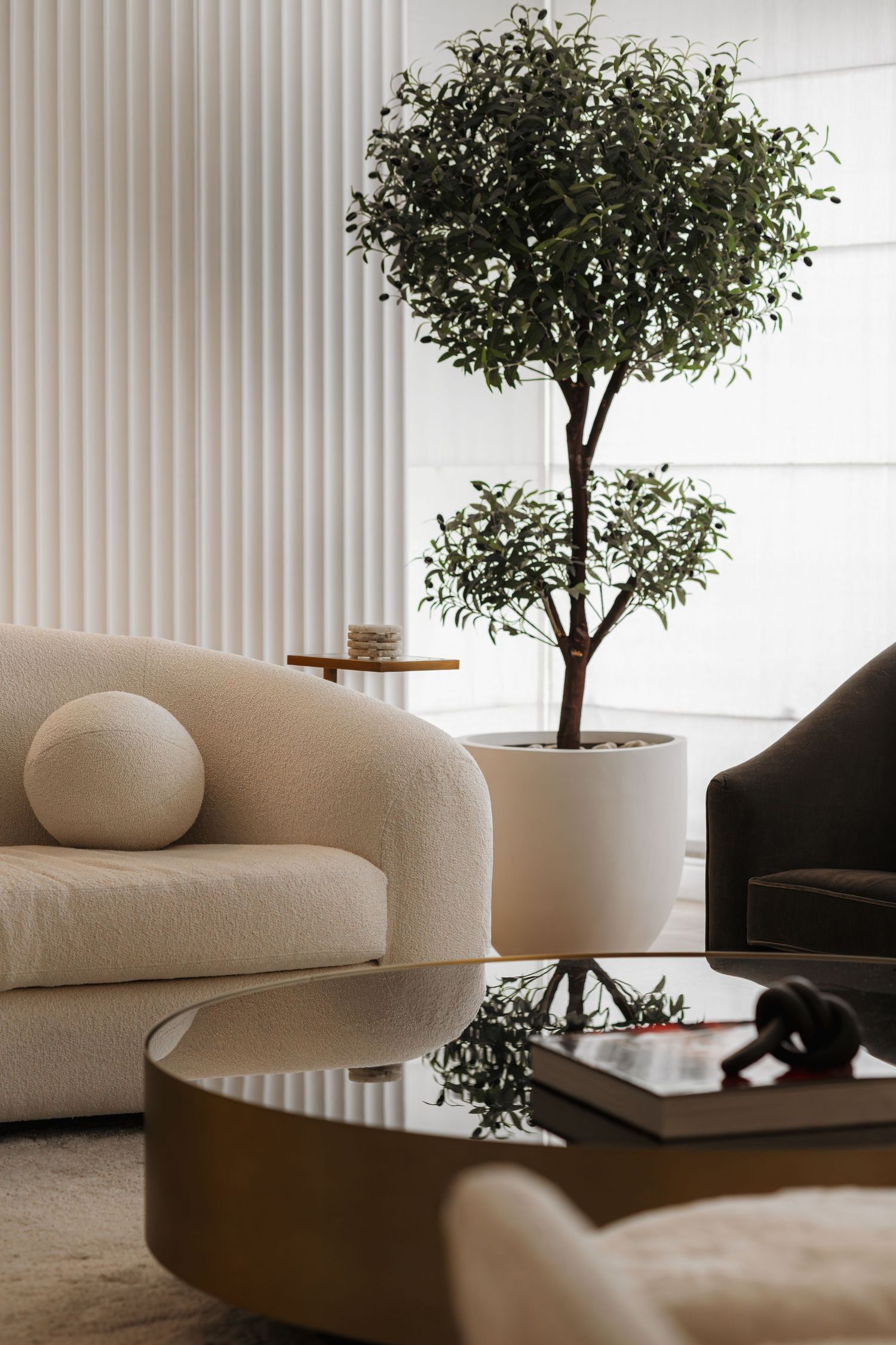 Modern living room detail with white bouclé sofa, large potted olive tree, gold glass coffee table, and dark armchair.