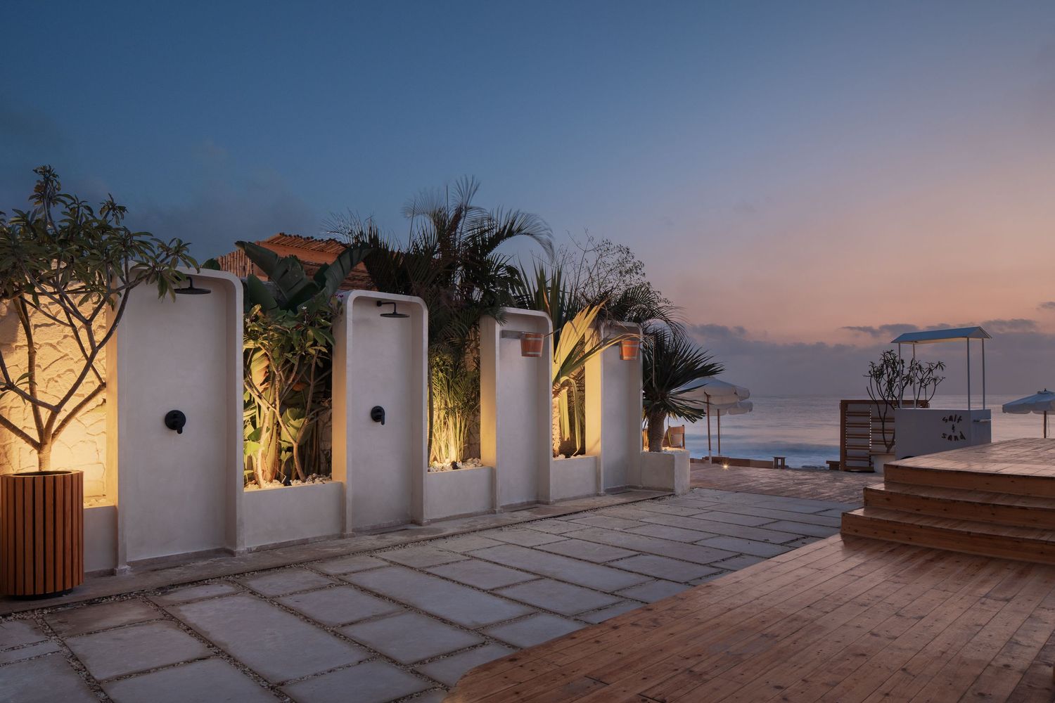Coastal outdoor terrace at dusk with white privacy screens, tropical palms, warm lighting, and ocean horizon view.