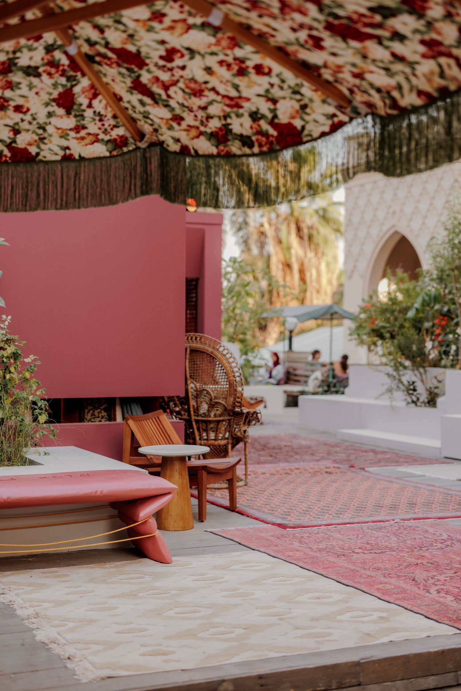 Bohemian outdoor seating area with floral canopy, wicker chair, pink wall, and vintage rugs.