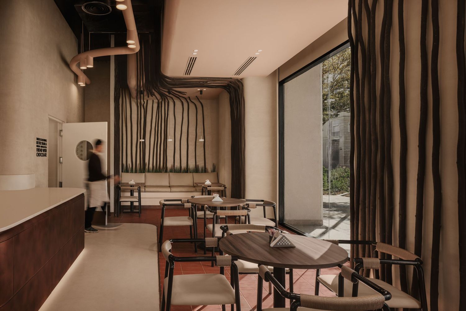 Moody restaurant interior with organic branch-like wall decor, dark wood furniture, and large window view.