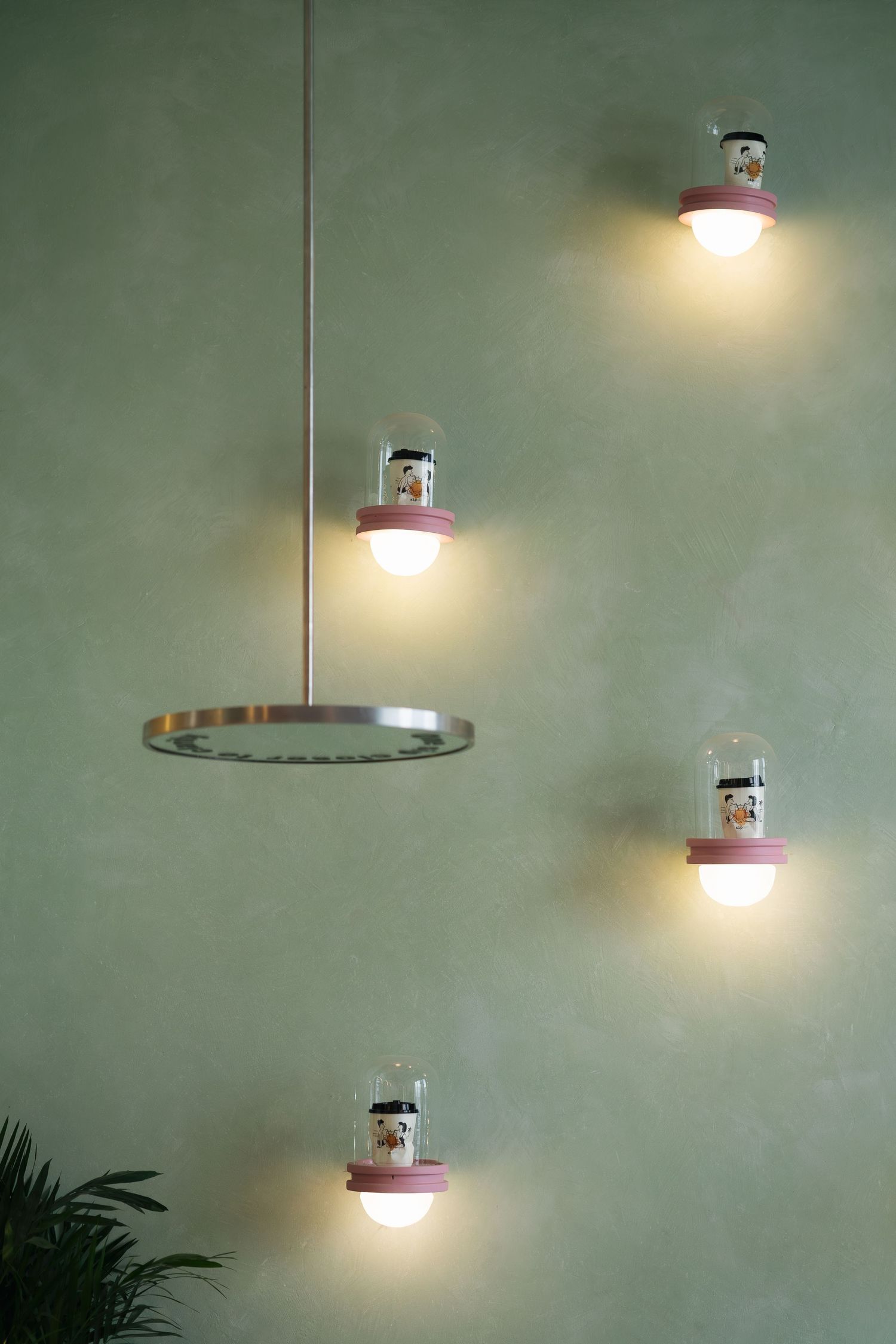 Modern wall-mounted and pendant lights with pink accents and snow globe details glowing warmly on a sage green wall.