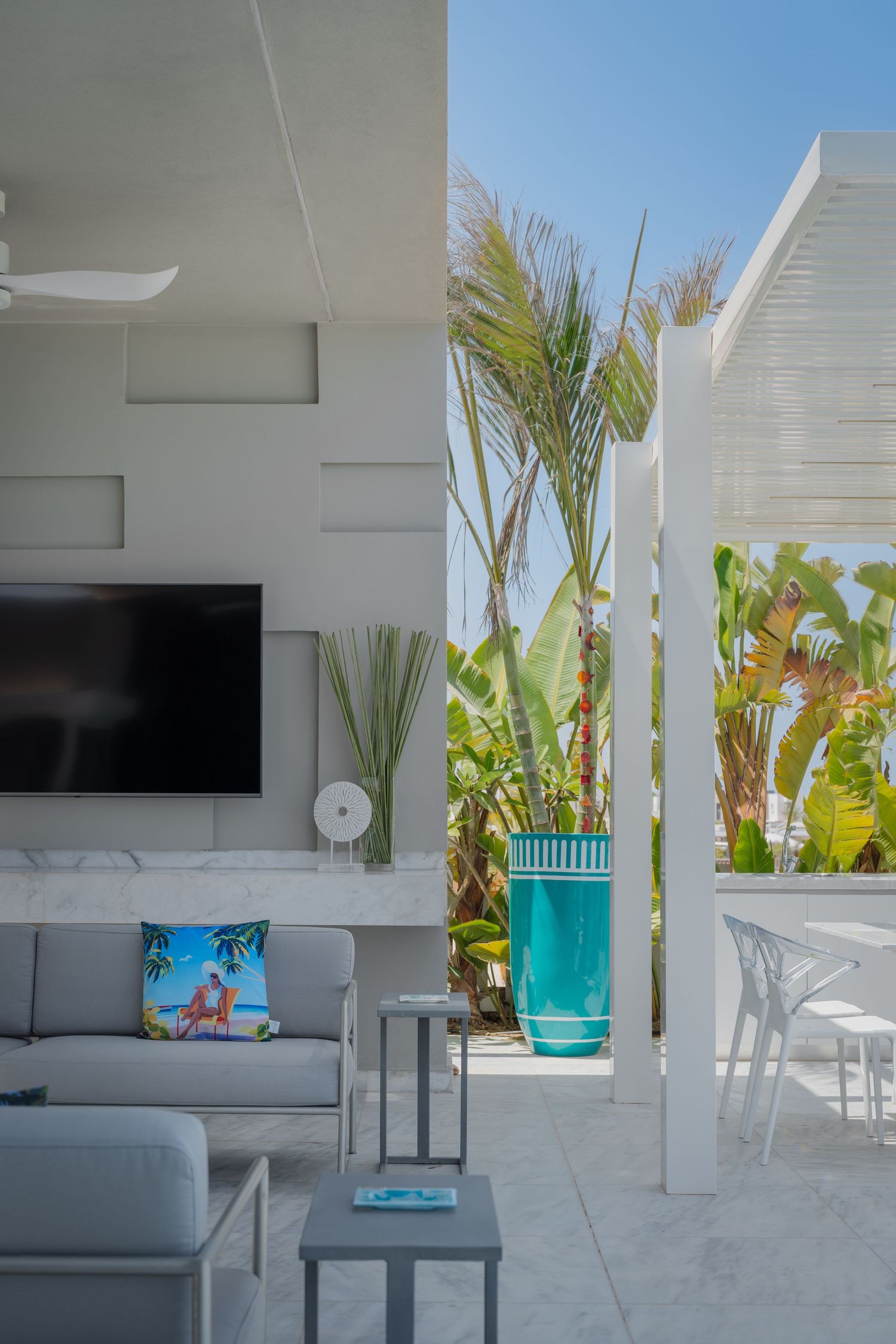 Contemporary outdoor lounge with gray sofa, wall-mounted TV, tall turquoise vase, and tropical palm trees.