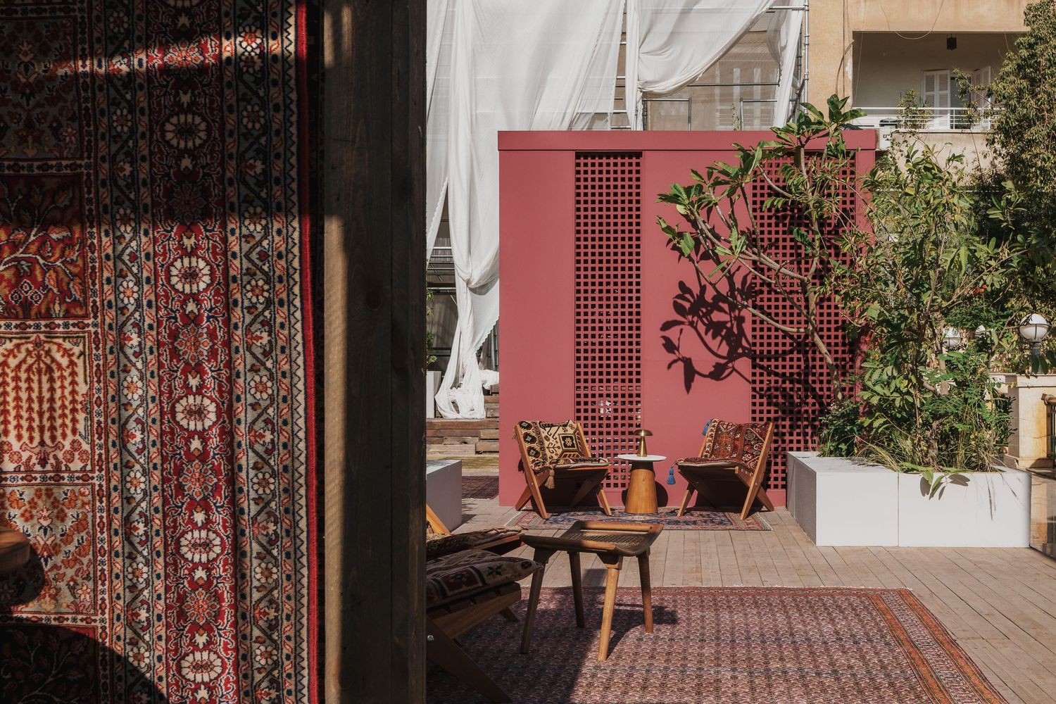 Bohemian rooftop terrace with Persian rugs, wooden chairs, and a pink lattice screen surrounded by lush plants.