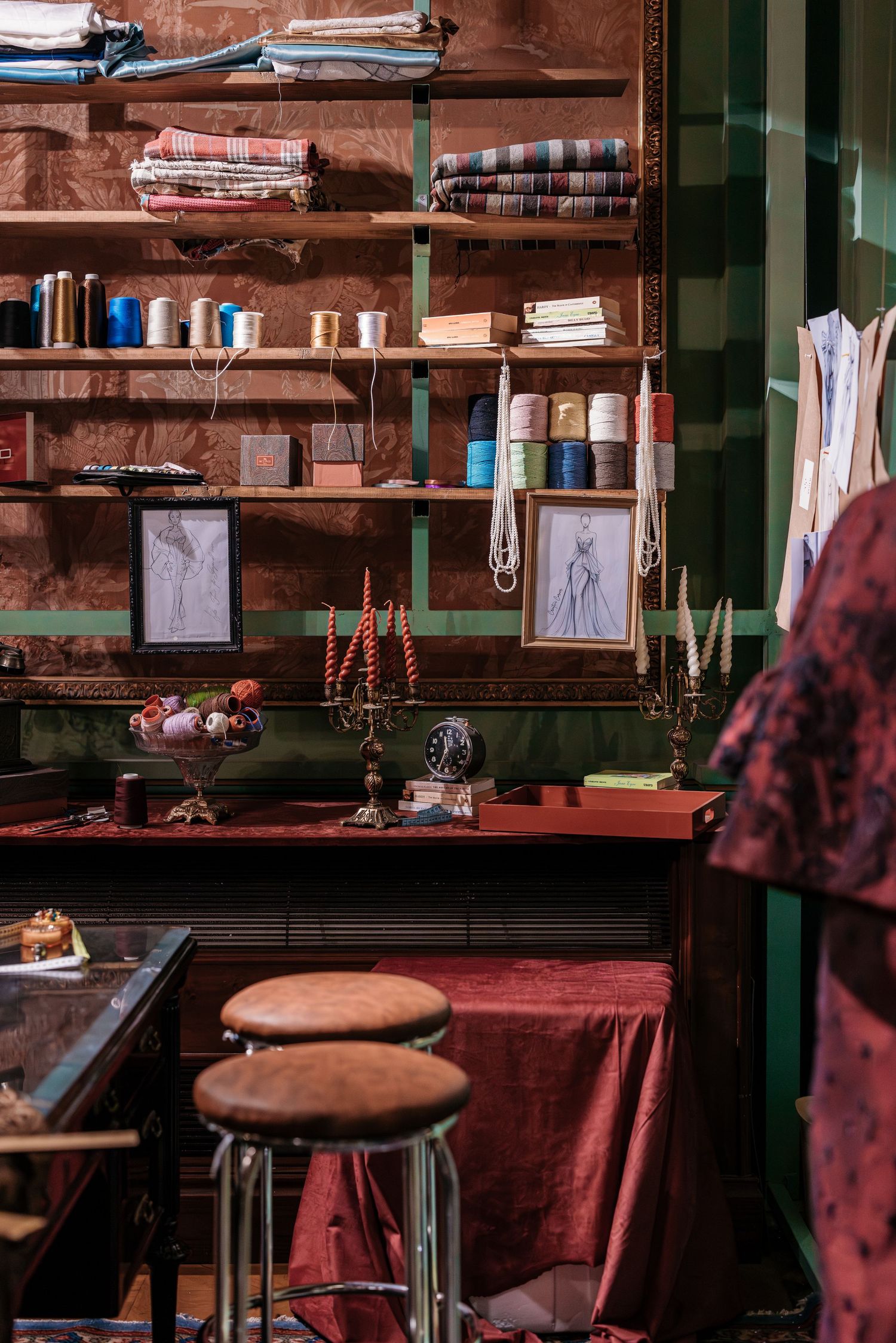 Vintage tailor workshop with thread spools, fabric rolls, fashion sketches, and red velvet work table.