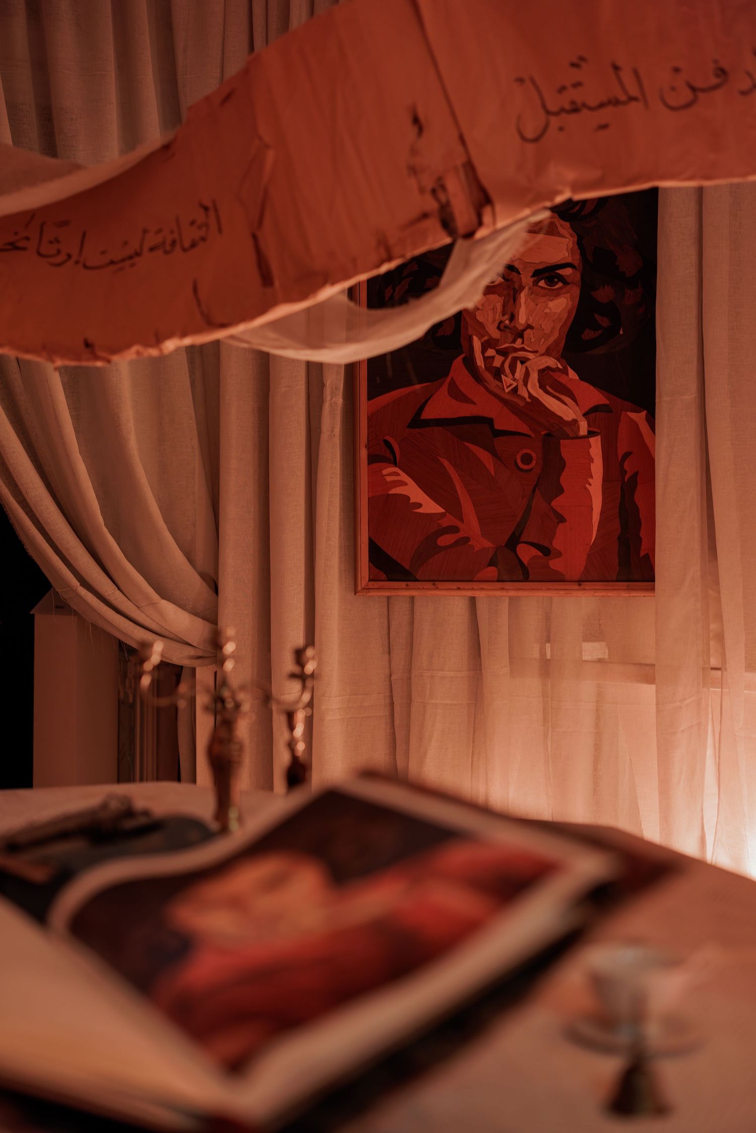 Open art book and painting of a pensive woman displayed in a warm red-lit room with Arabic text banners.