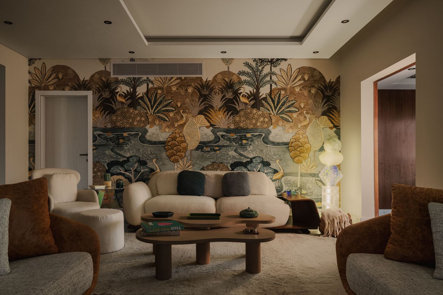 Elegant living room with gold tropical botanical wallpaper mural, curved cream sofa, rust armchairs, and round coffee table.