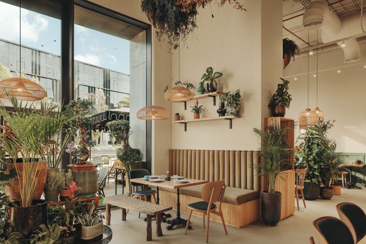 Bright cafe interior with natural wood furniture, wicker pendant lights, and lush tropical plants throughout.