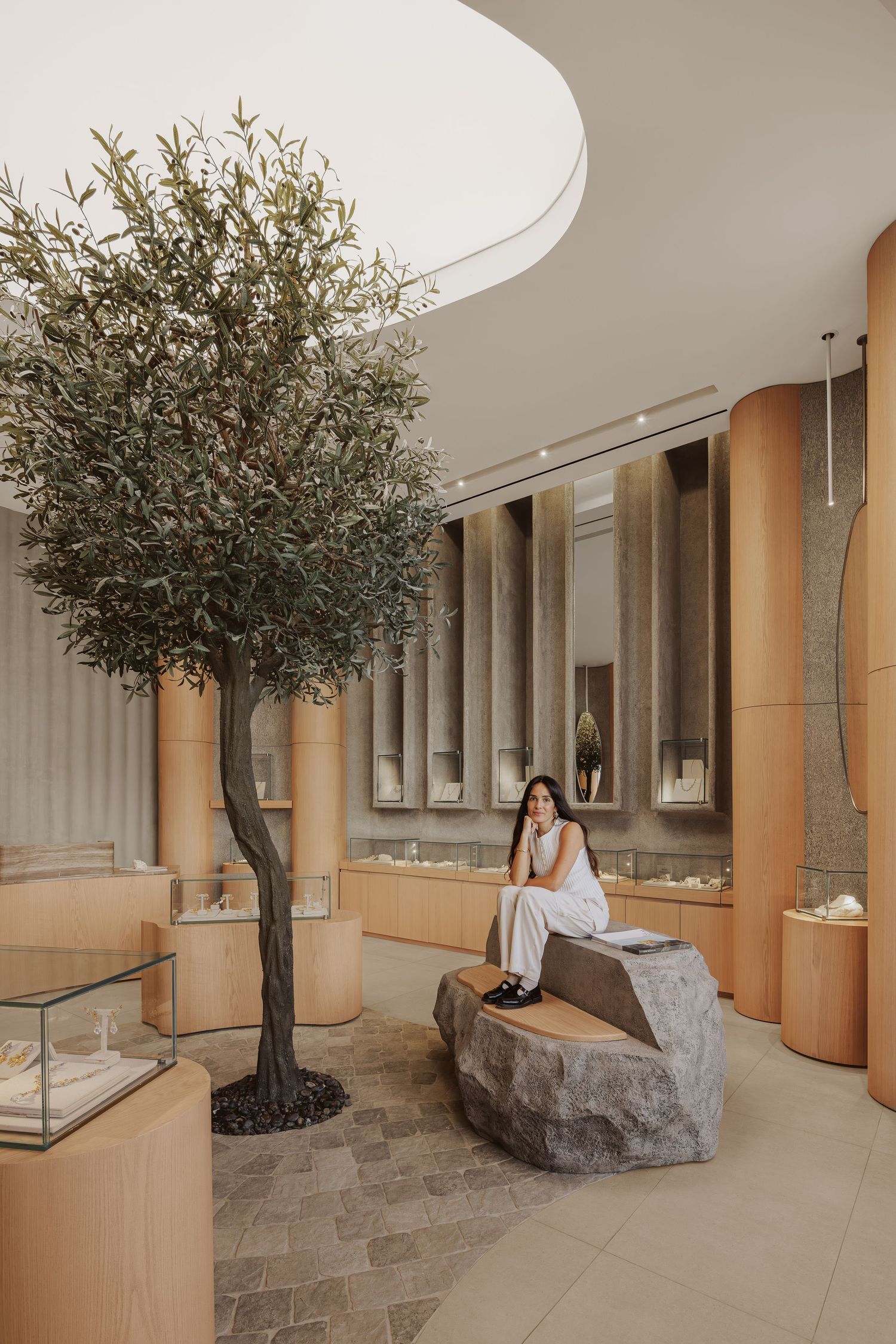 Woman in white outfit sitting on stone bench beside olive tree in elegant jewelry store interior.