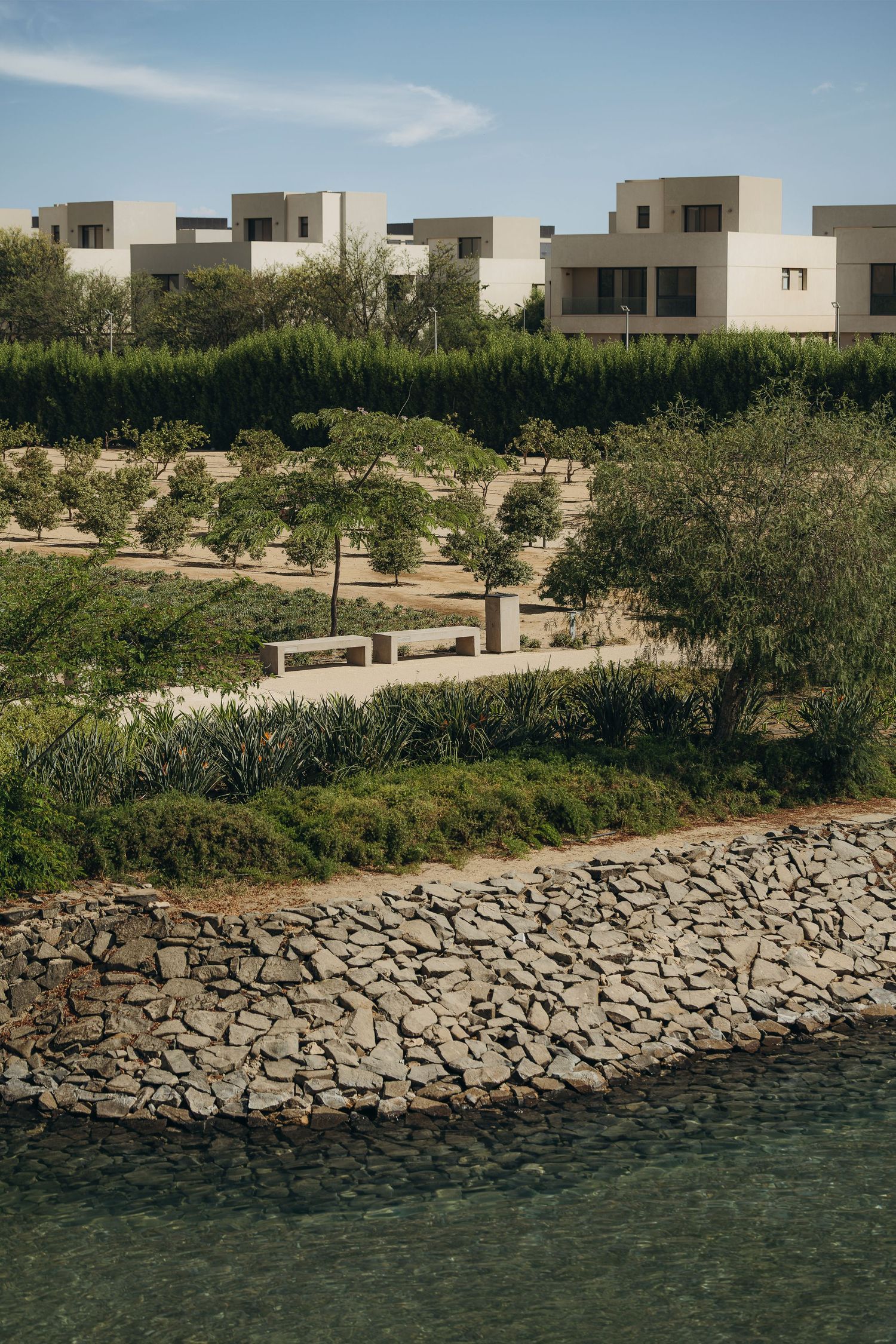Stone-edged waterway beside lush gardens and modern white cubic villas under a clear blue sky.