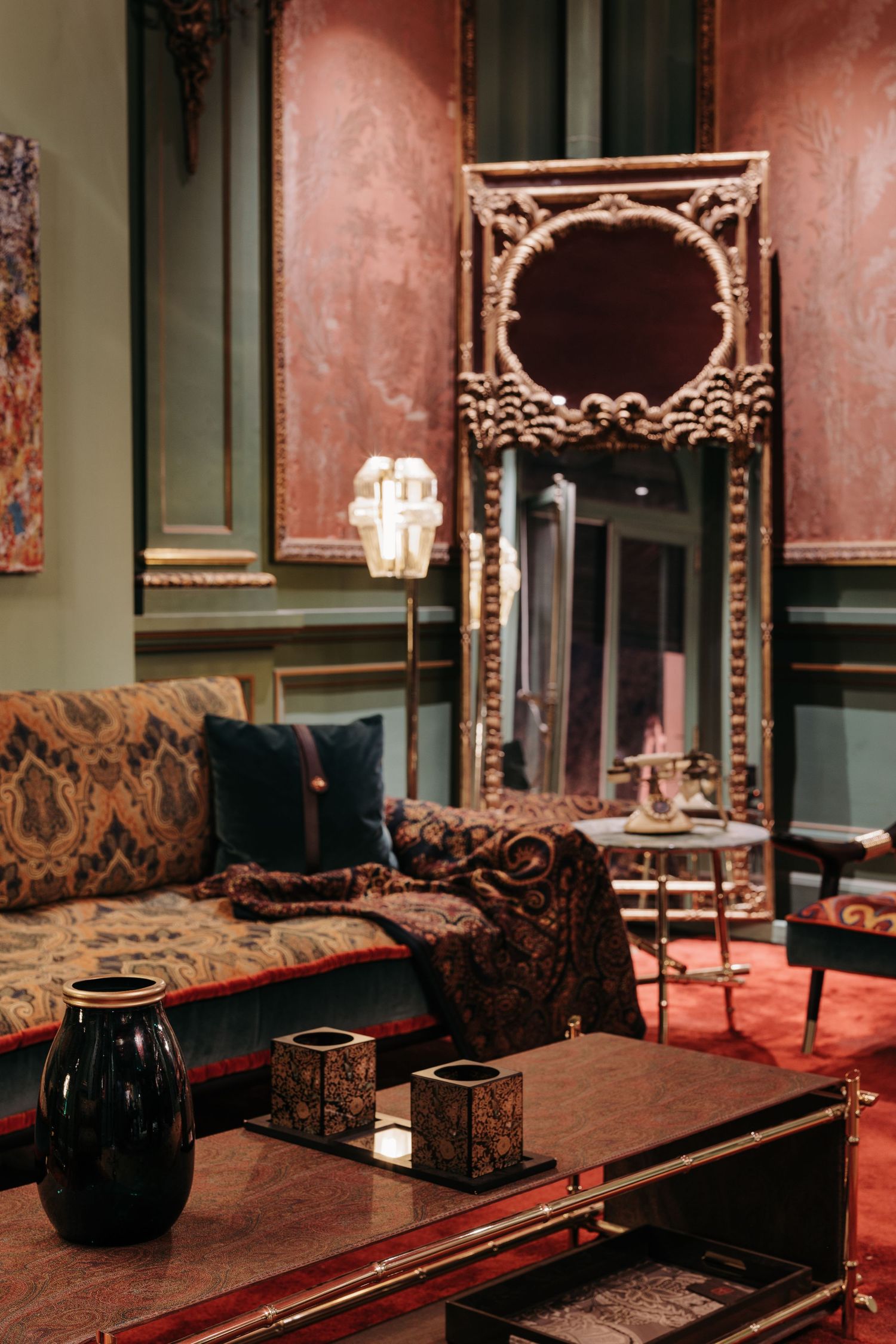 Opulent lounge with ornate gold mirror, tapestry sofa, velvet cushions, and decorative vases on table.
