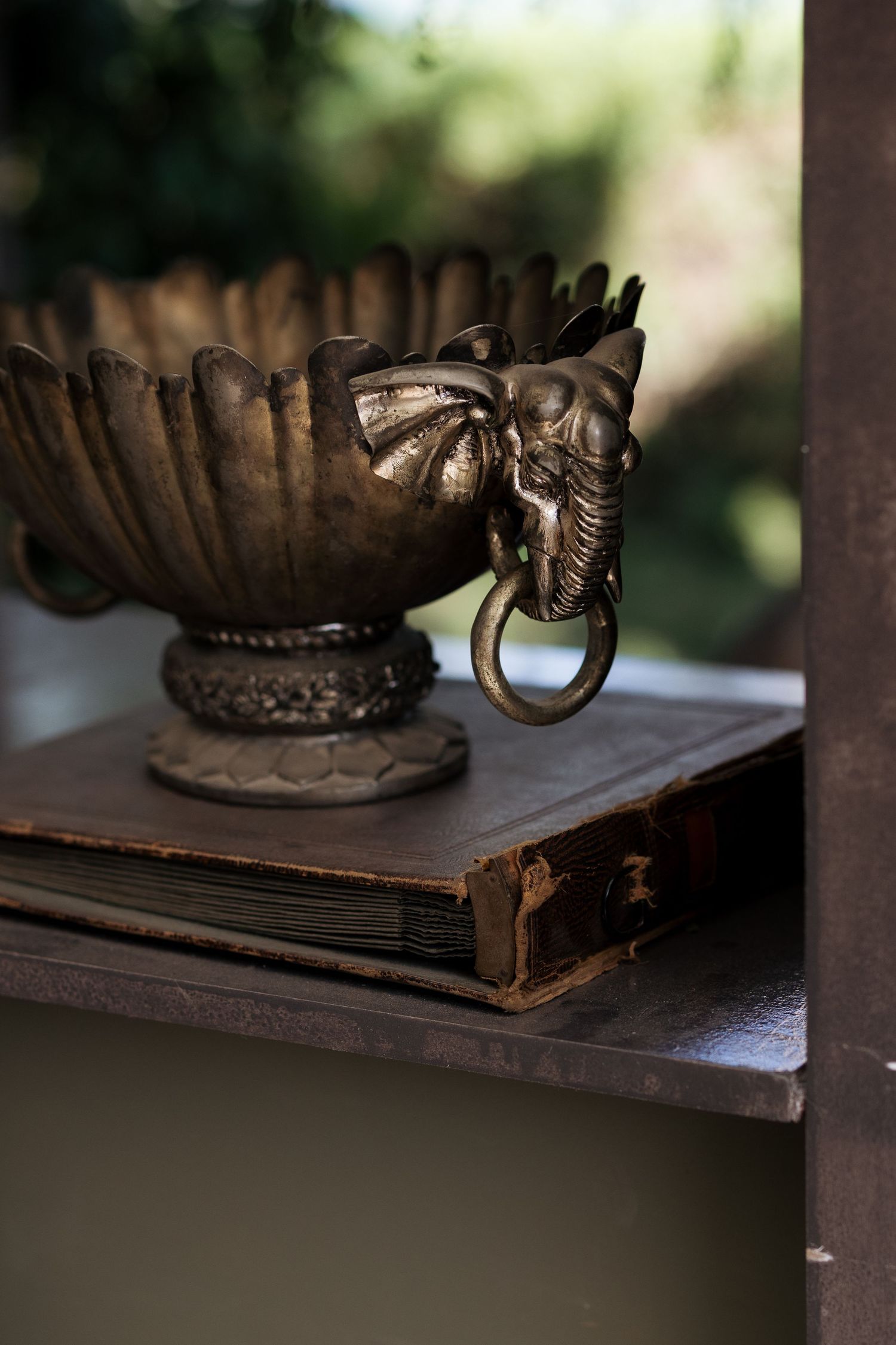 Ornate bronze elephant-handled bowl atop an aged leather book on a dark wooden shelf.