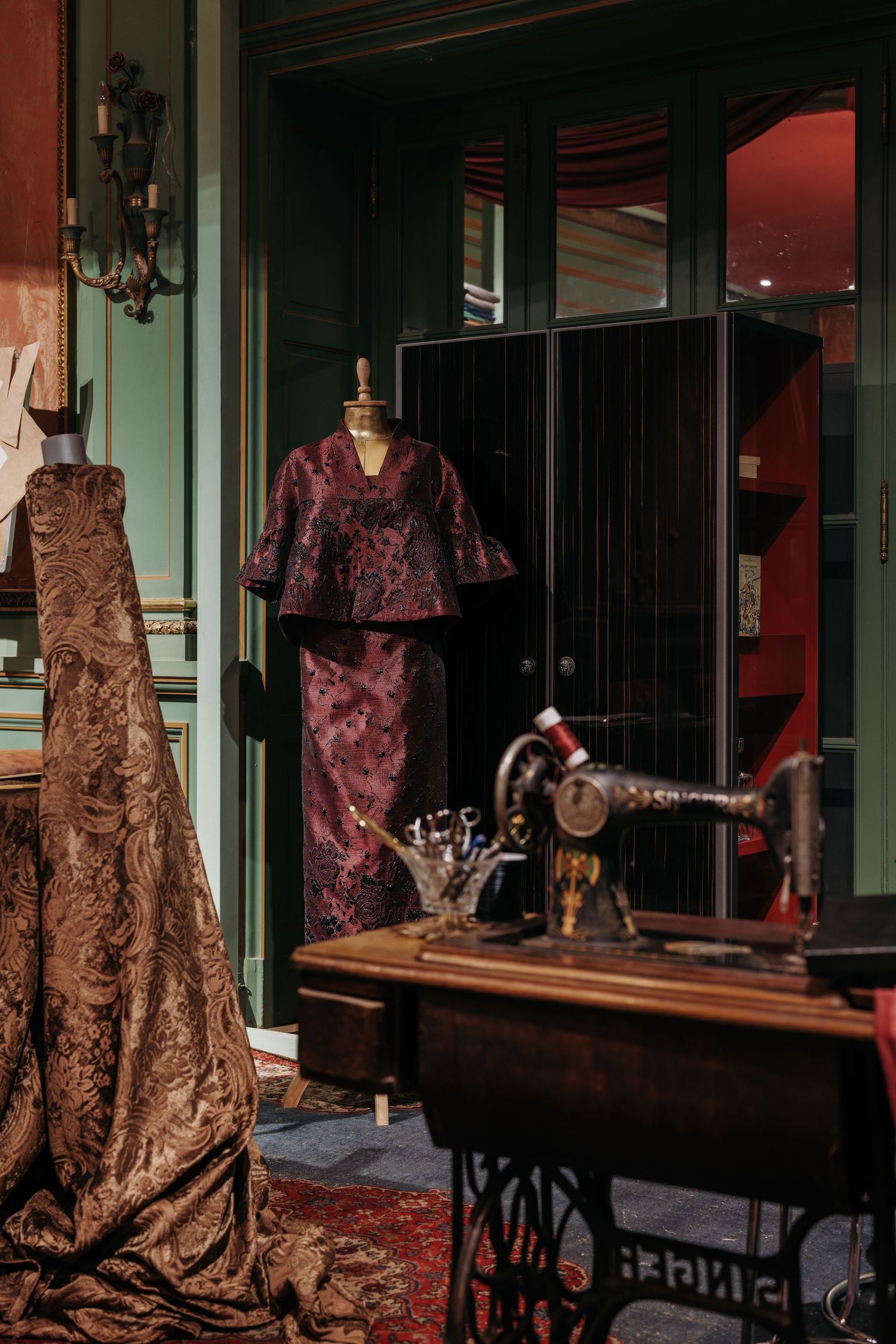 Luxury fashion atelier featuring vintage sewing machine, bronze brocade gown, and burgundy patterned suit on mannequin.