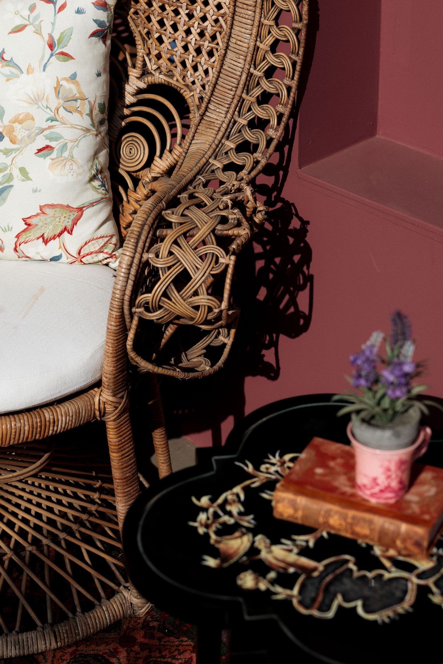 Ornate wicker peacock chair with floral cushion beside a black table with books and lavender.