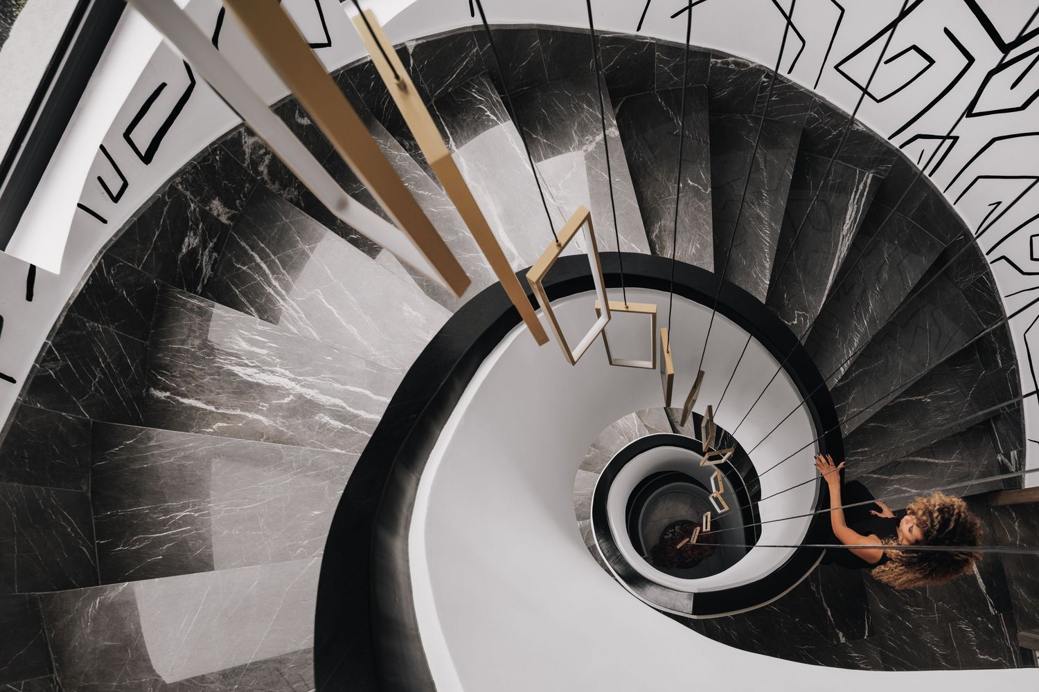 Overhead view of spiral marble staircase with gold geometric railing and person descending, framed by graphic wall mural.
