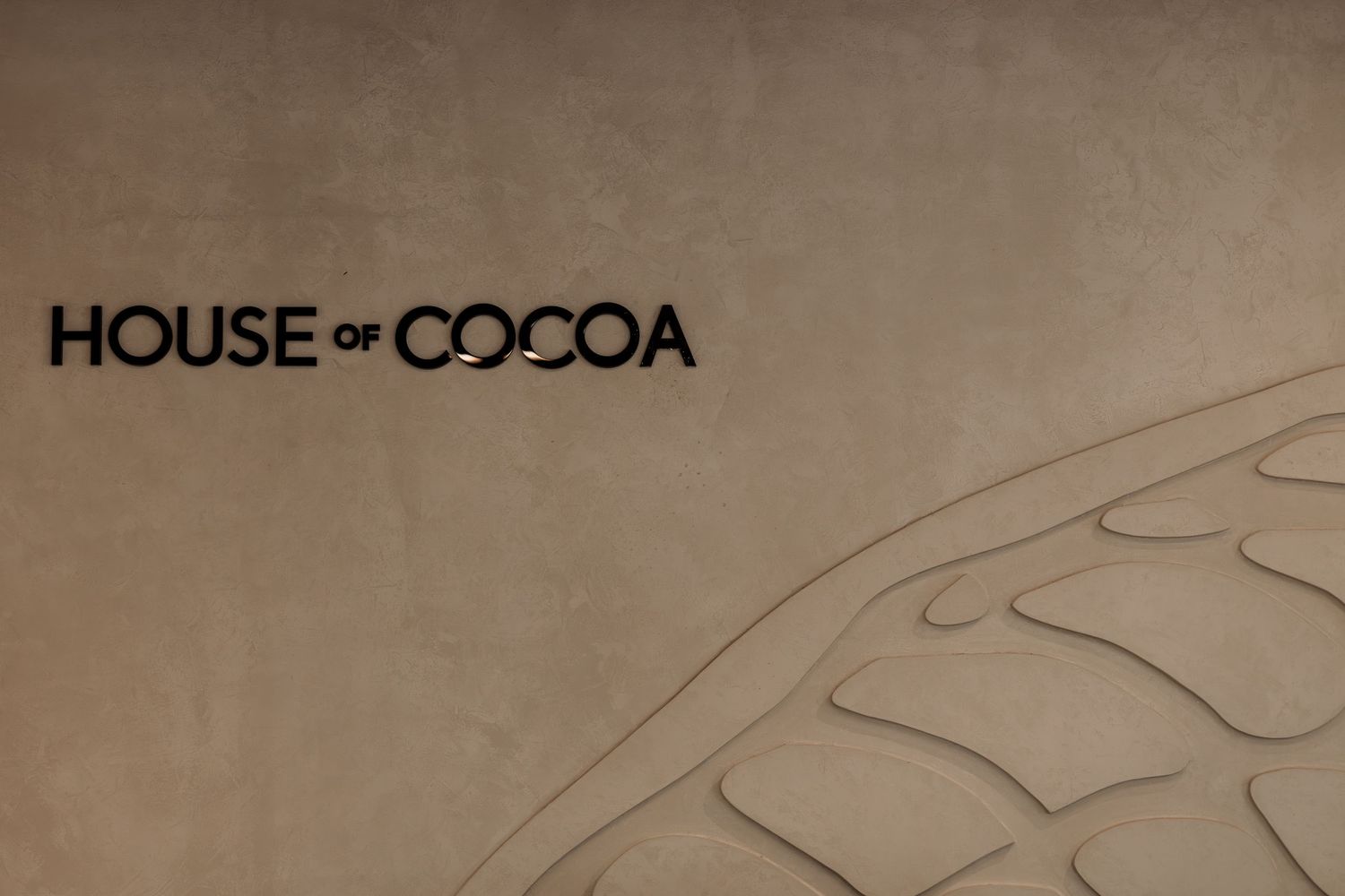 House of Cocoa logo on a textured beige wall with subtle cocoa pod relief design.
