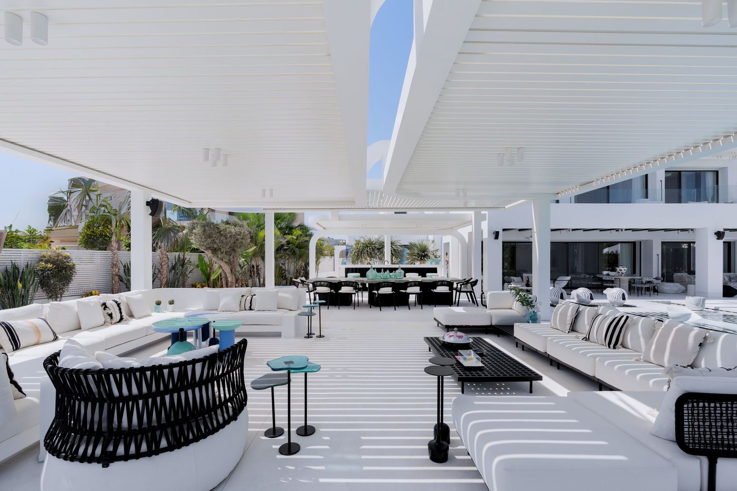 Spacious white luxury outdoor lounge with circular sofas, striped flooring, pergola ceiling, and lush garden backdrop.