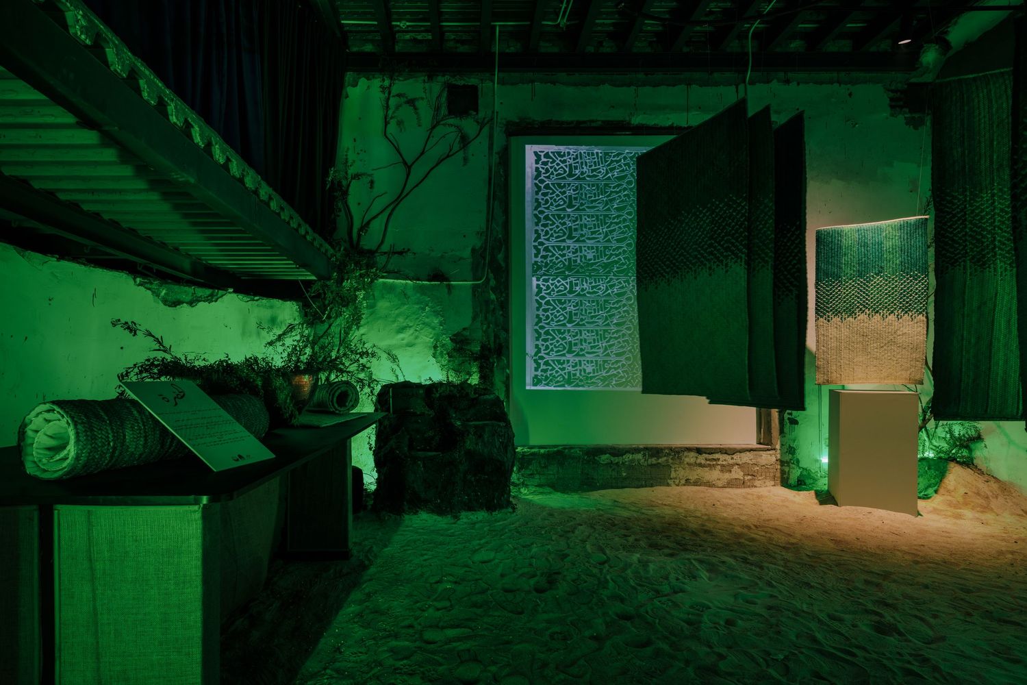 Green-lit atmospheric room with sand floor, plants, and glowing window art installation.