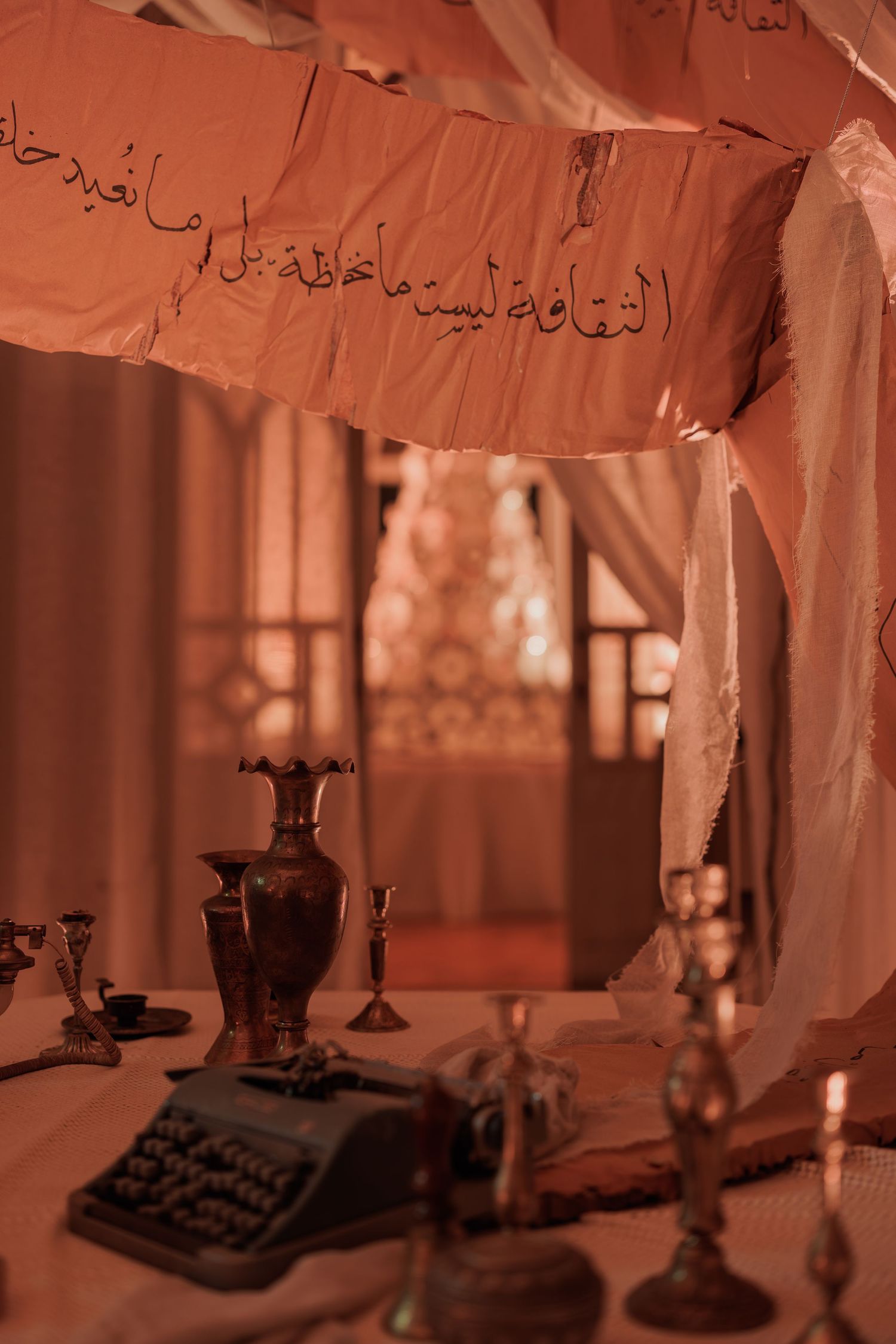 Vintage typewriter and brass candlesticks on a table beneath hanging Arabic calligraphy paper in warm red light.