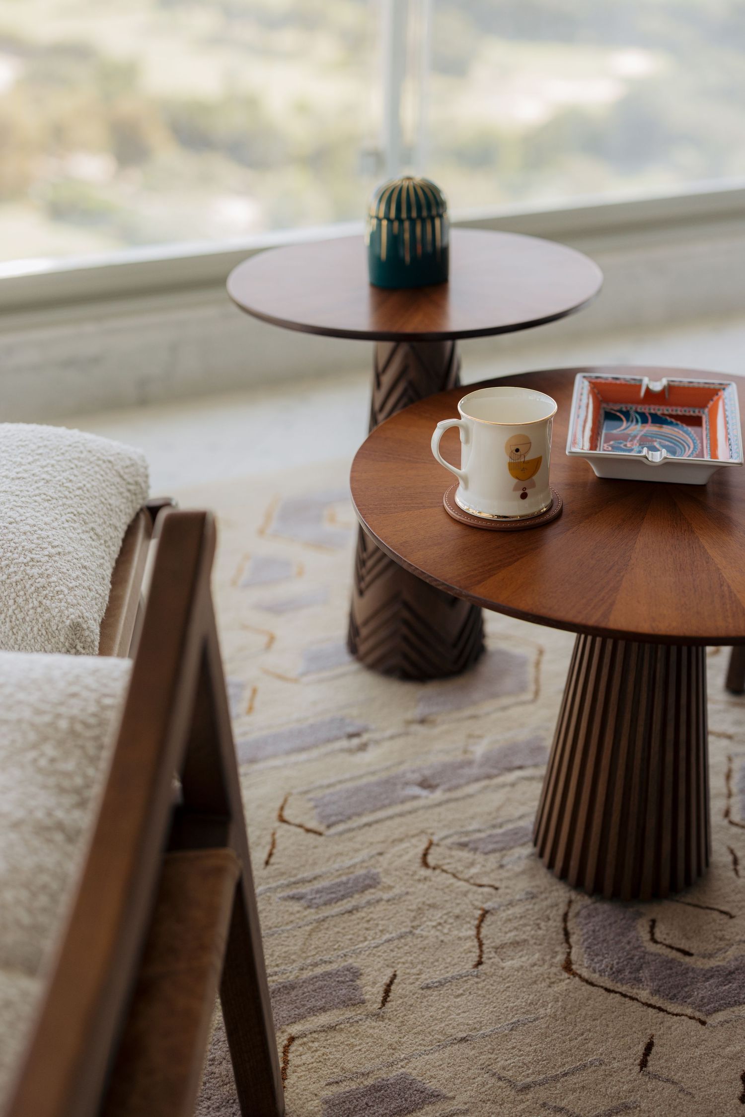 Mid-century modern wooden side tables with ribbed pedestals, ceramic mug, and cactus near large floor-to-ceiling window.