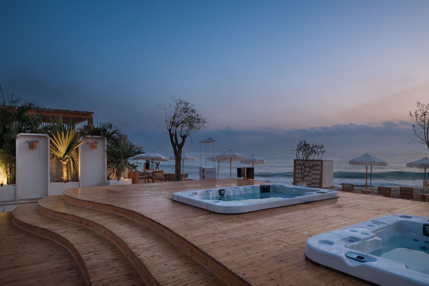 Upscale seaside terrace at dusk with two jacuzzis, tiered wooden decking, white umbrellas, and panoramic sea views.