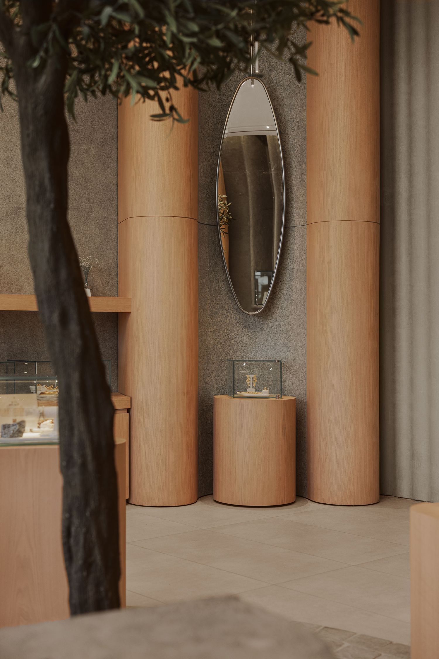 Minimalist jewelry store vignette with tall wood columns, oval hanging mirror, and cylindrical display pedestal.