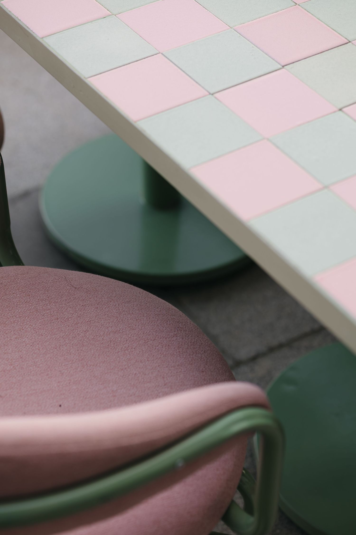 Detail of green metal chair and pink cushion beside a checkerboard tiled table with green pedestal base.