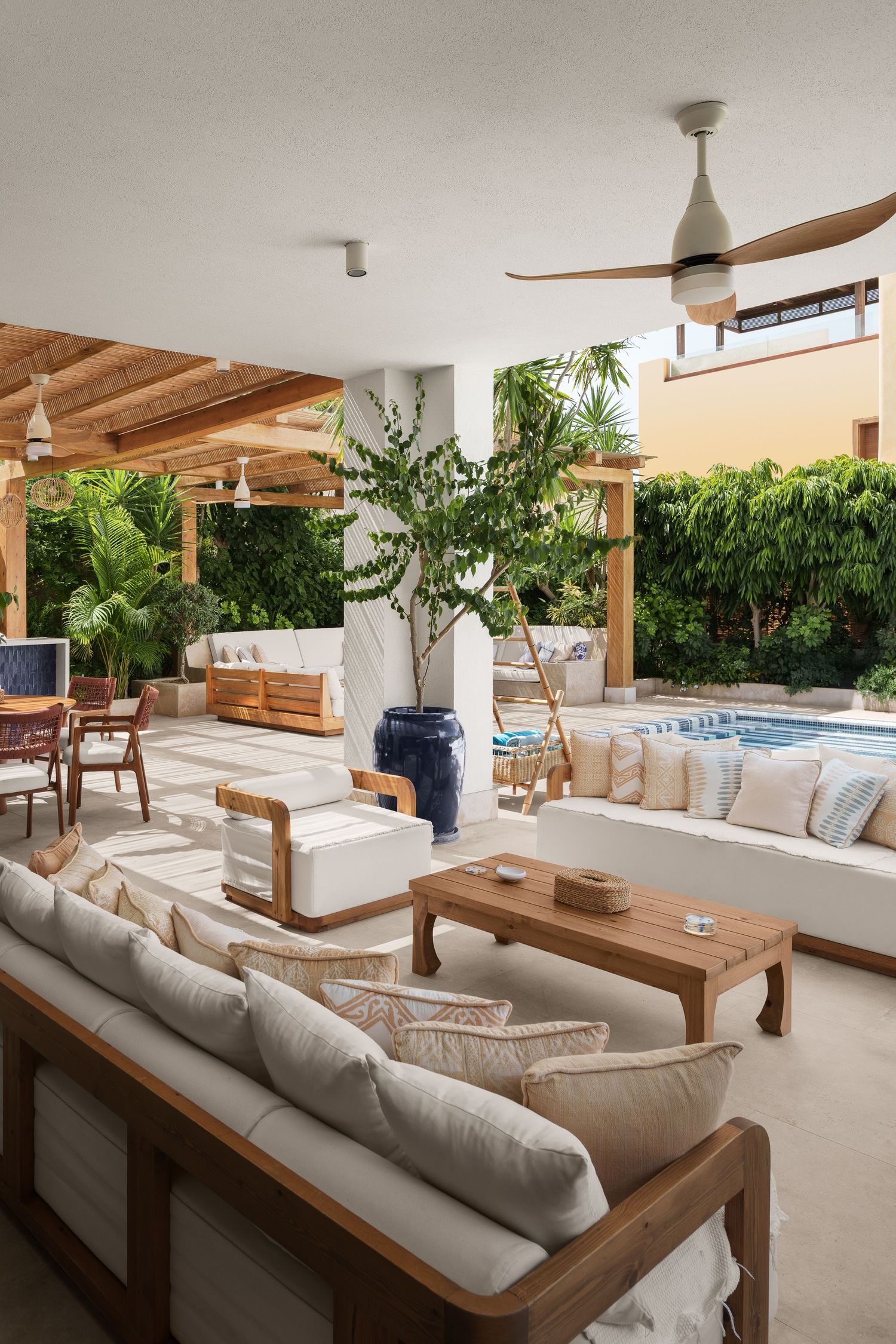 Luxury open-air living space with white sofas, wooden pergola, ceiling fan and tropical pool garden.