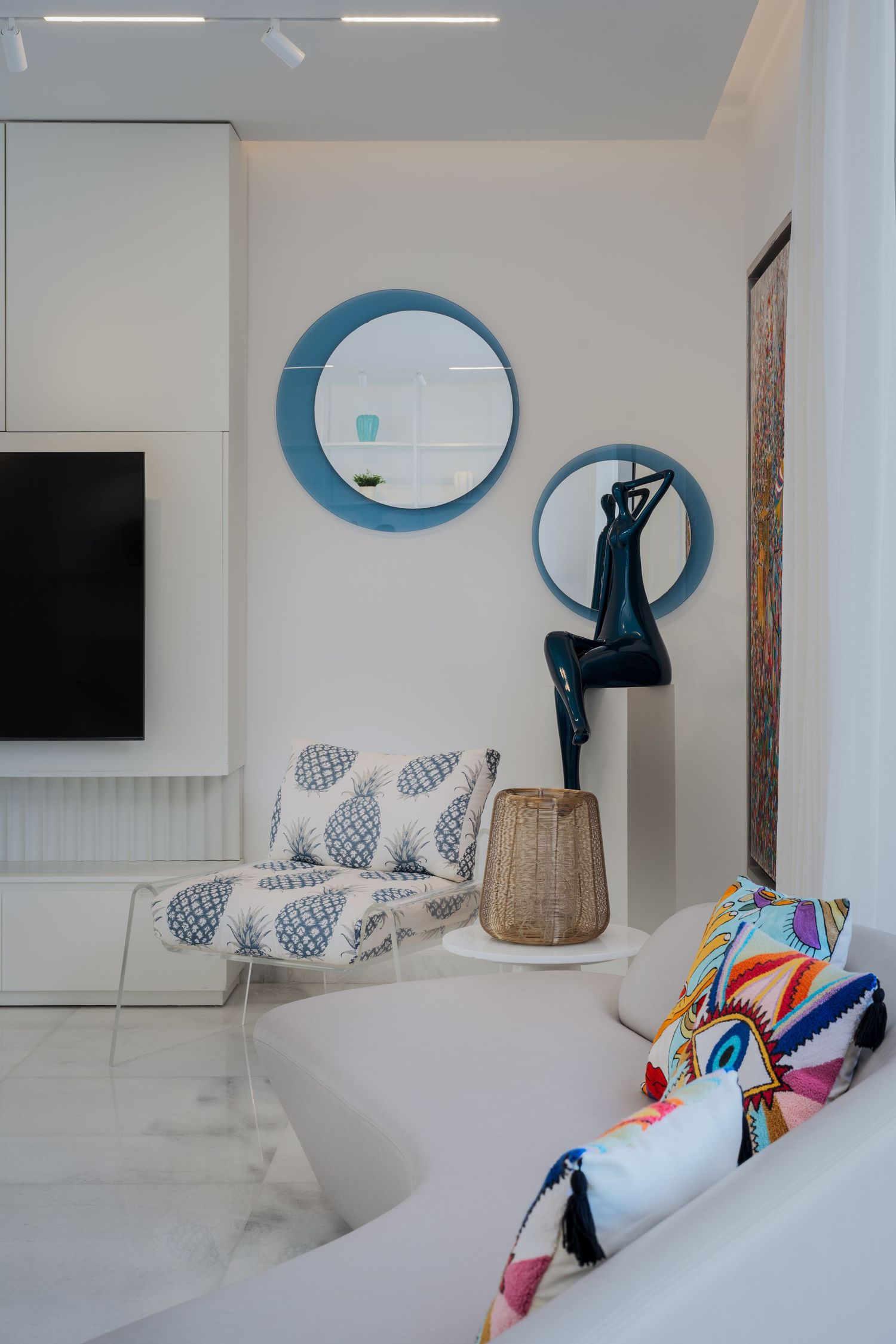 Modern white living room with blue circular mirrors, dark sculpture, patterned accent chair, and colorful throw pillows.