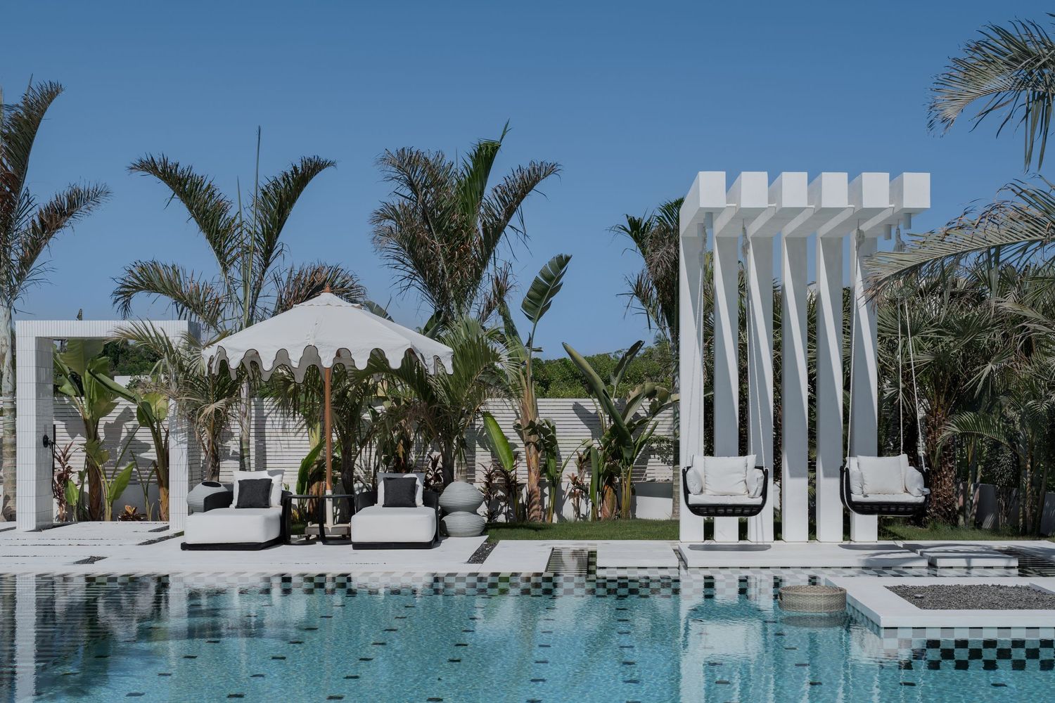 Resort pool with white lounge chairs, decorative pergola structures, tropical palm trees, and a striped cabana tent.