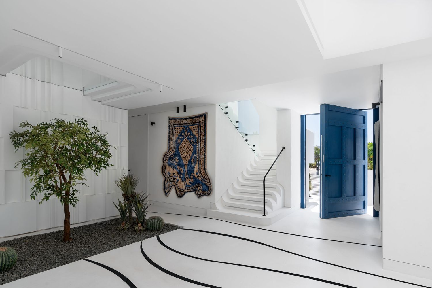 Elegant white villa entrance with indoor garden, decorative tapestry, curved staircase, and vivid blue door.