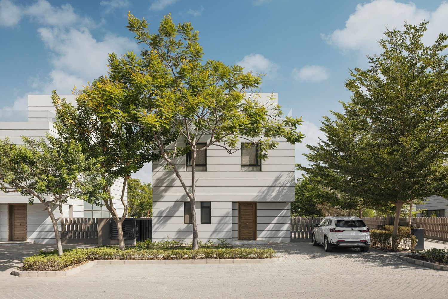 Modern white two-story house with lush green trees, wooden door accent, and parked white car in paved driveway.