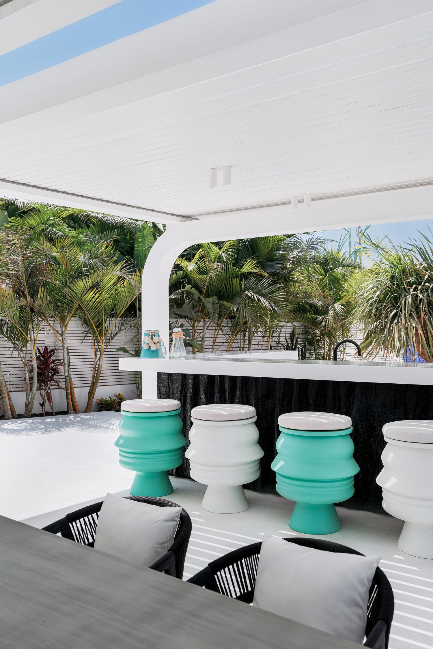 Outdoor bar area with teal and white barrel stools, black counter, white pergola, and lush tropical palms.
