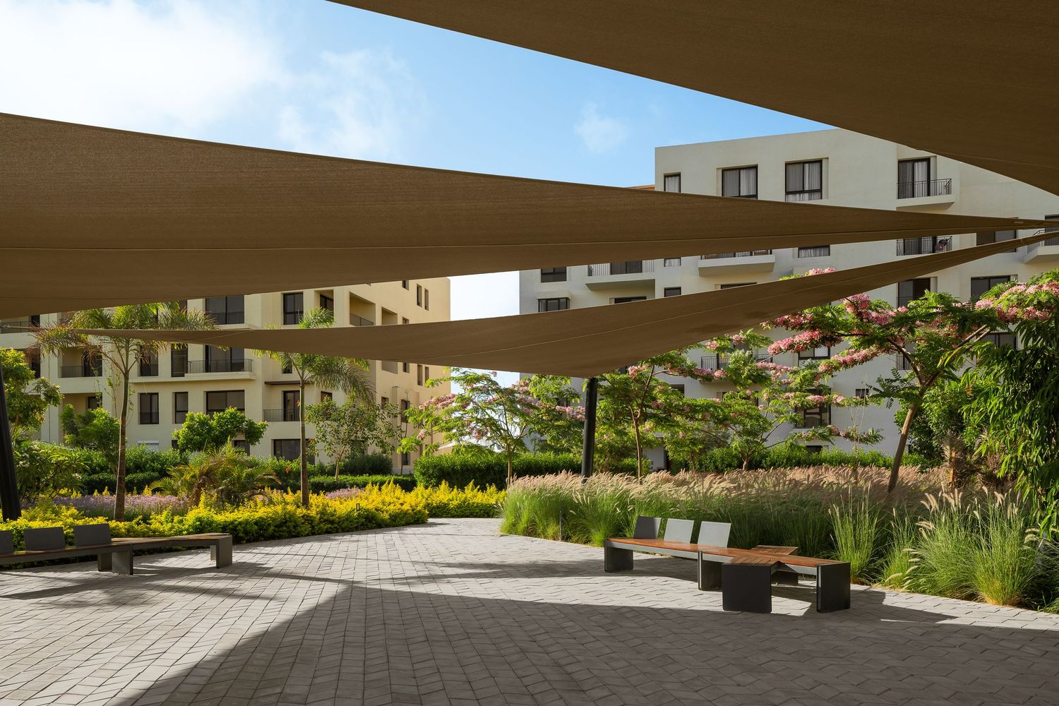 Spacious paved courtyard with large triangular shade sails, wooden benches, and colorful landscaped gardens.