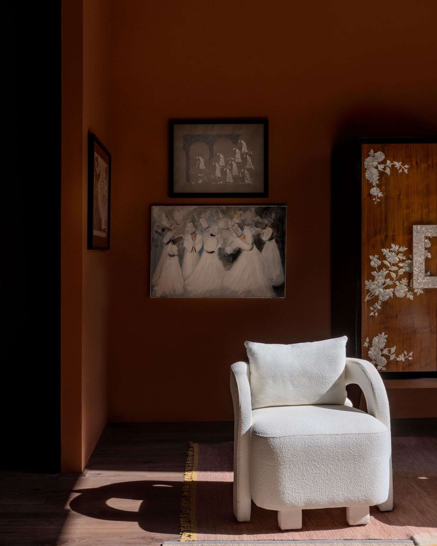White boucle armchair in warm terracotta room with vintage artwork and ornate wooden cabinet.