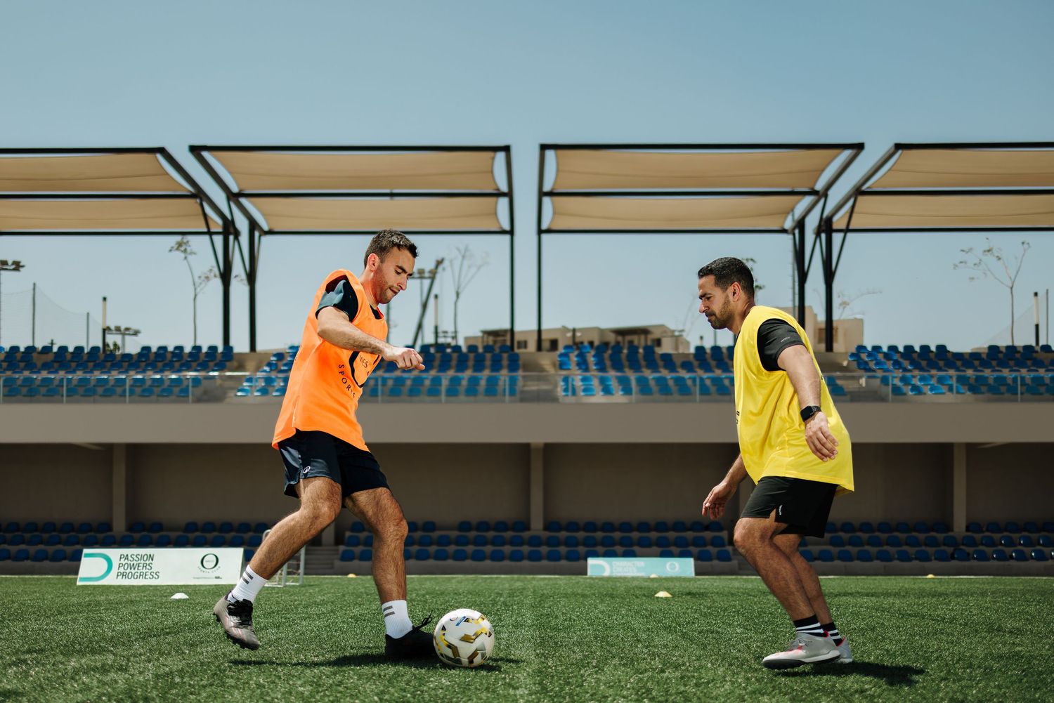 Two male soccer players in orange and yellow bibs practicing ball control on an artificial turf field.
