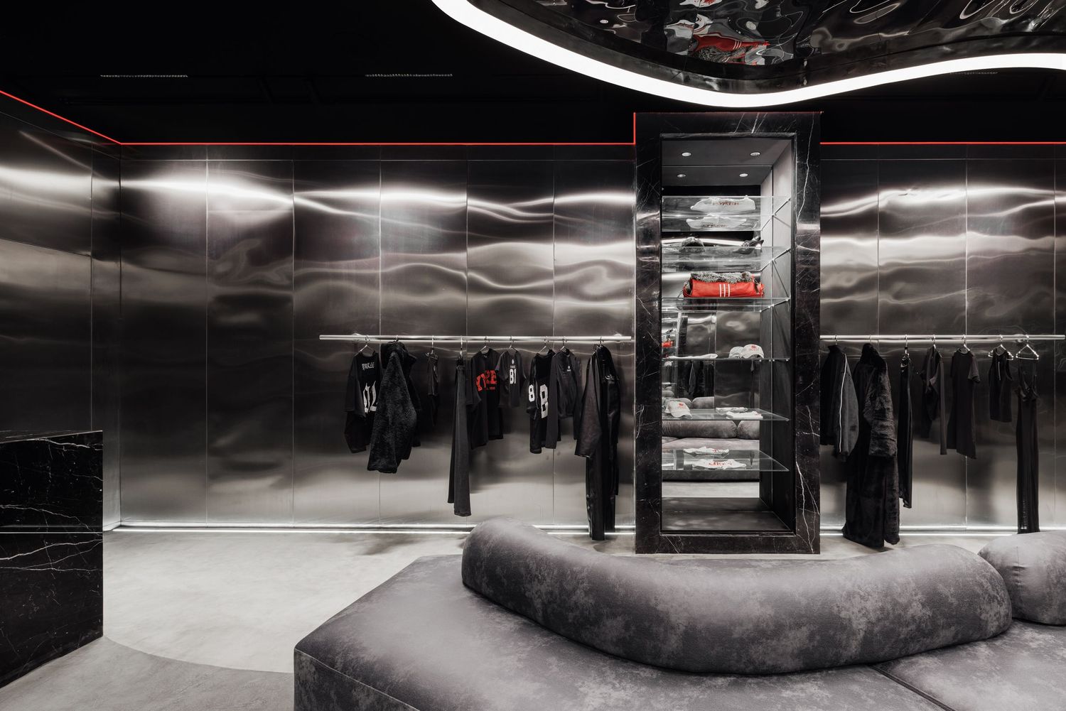 Upscale dark boutique interior with circular gray velvet seating, metallic walls, and black clothing racks under red accent lights.