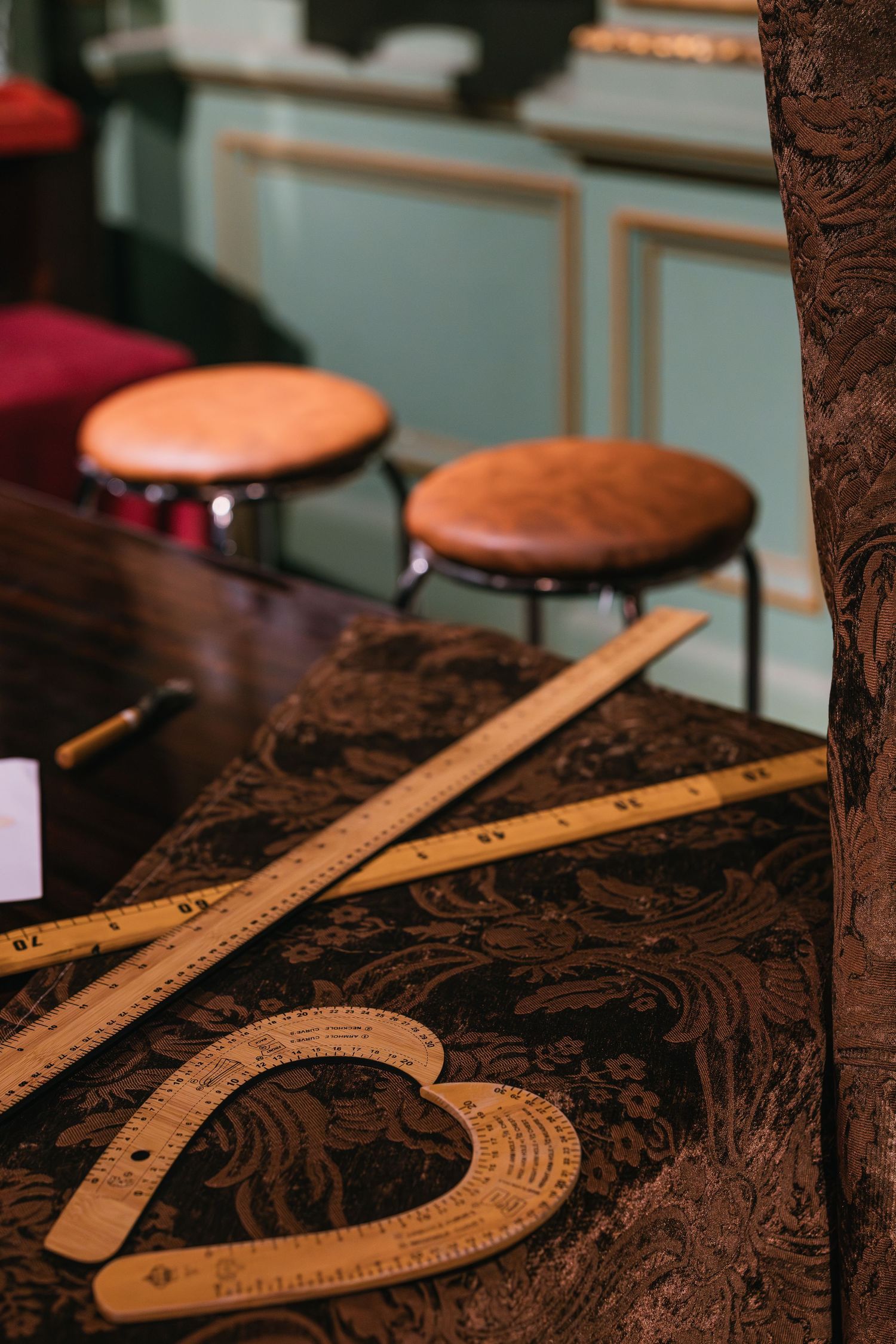Wooden rulers and a French curve on a dark ornate table with leather stools in background.