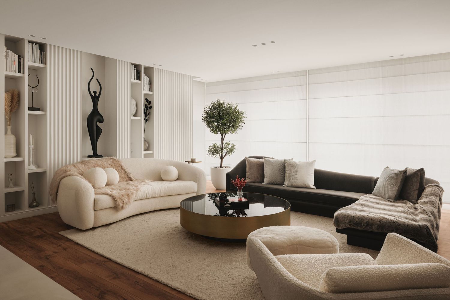 Elegant living room with curved beige sofas, black sectional, gold coffee table, indoor tree, and abstract black sculpture.
