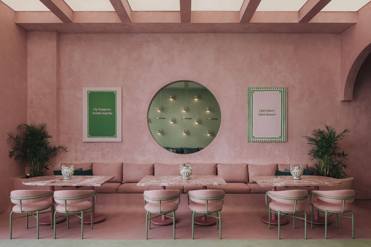 All-pink dining room with tufted circular mirror, framed art, banquette seating, and round marble tables.
