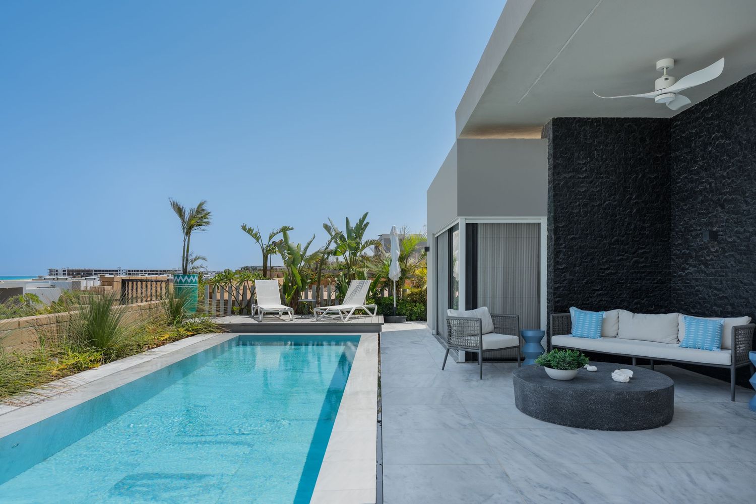 Luxury villa pool terrace with marble tiles, outdoor sofa, dark stone wall, and ocean horizon beyond.