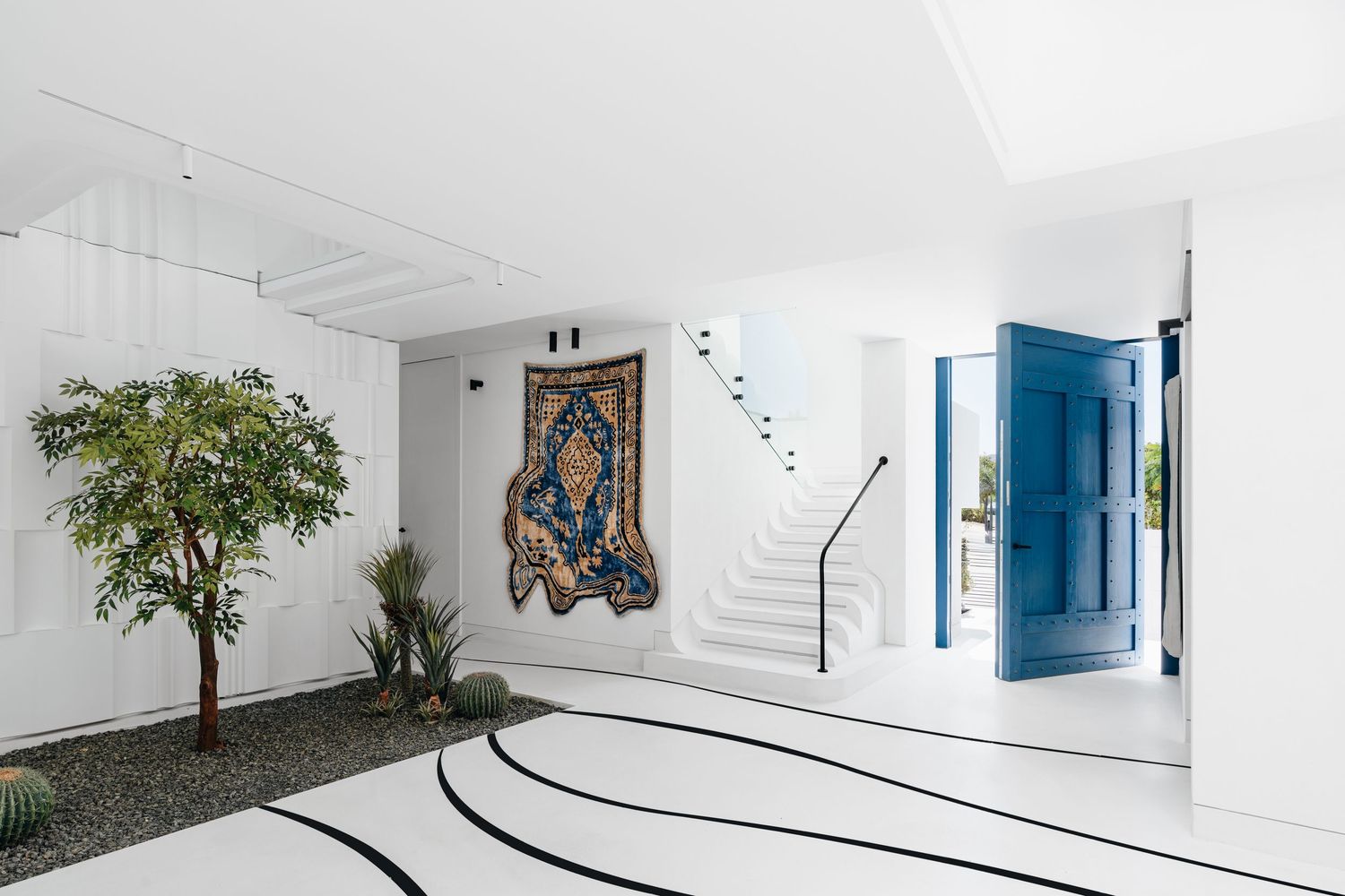 Modern white foyer with blue door, ornate wall tapestry, indoor tree, and curved black floor lines.