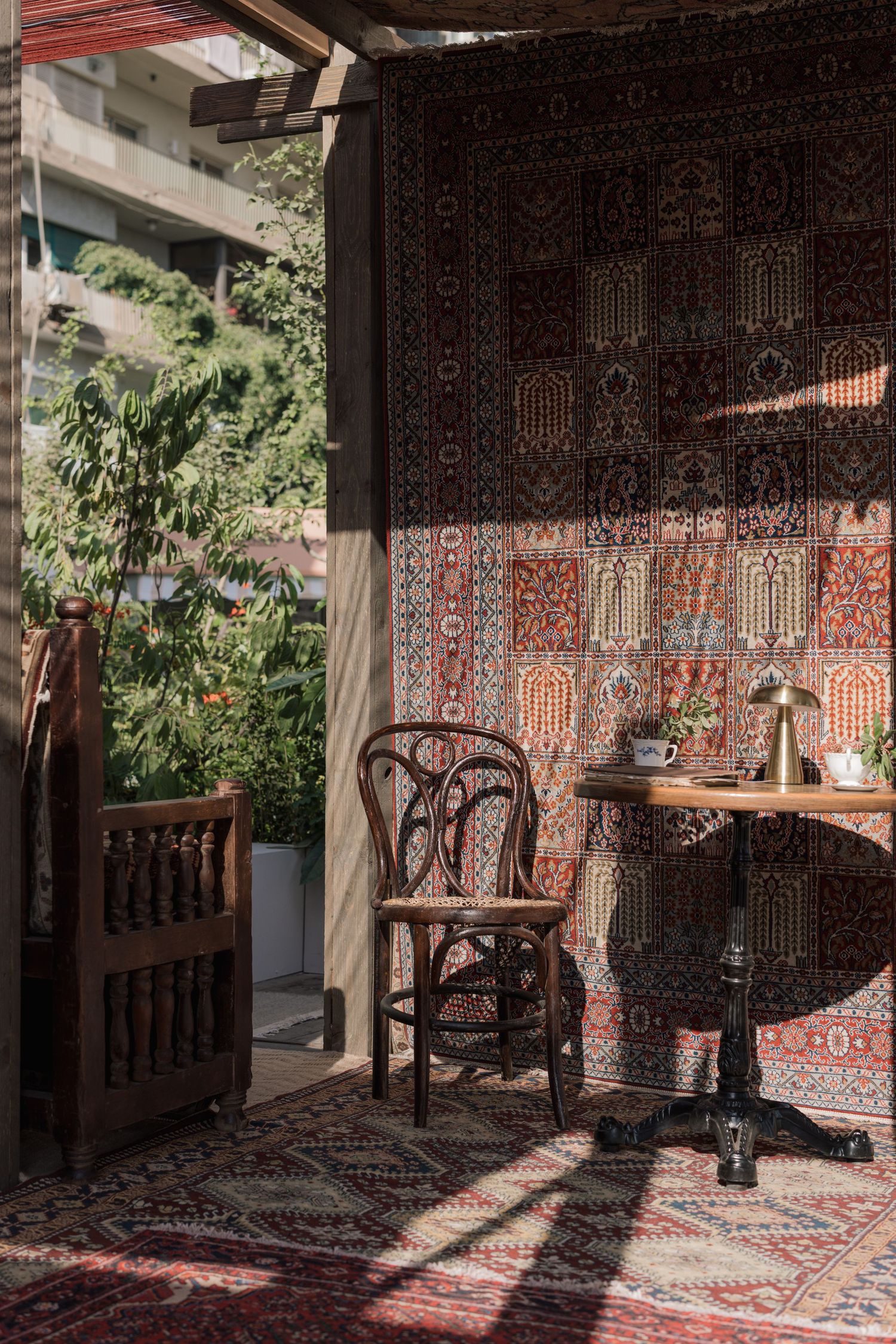 Sunlit balcony cafe with ornate Persian rug wall, Thonet chair, round table, and lush garden view.