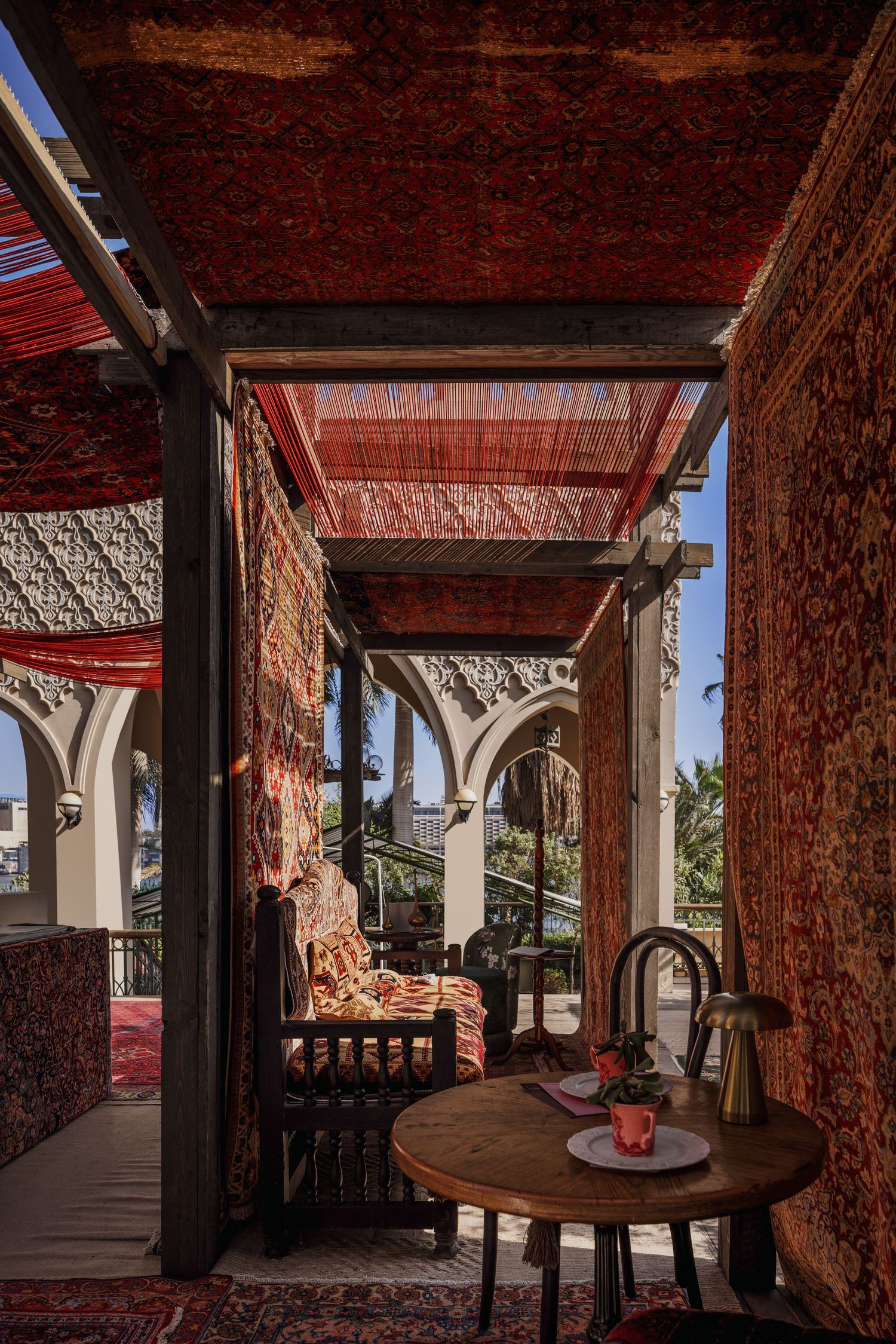 Ornate Middle Eastern terrace with red Persian rugs on ceiling and walls, wooden table with brass teapot, and arched stone details.