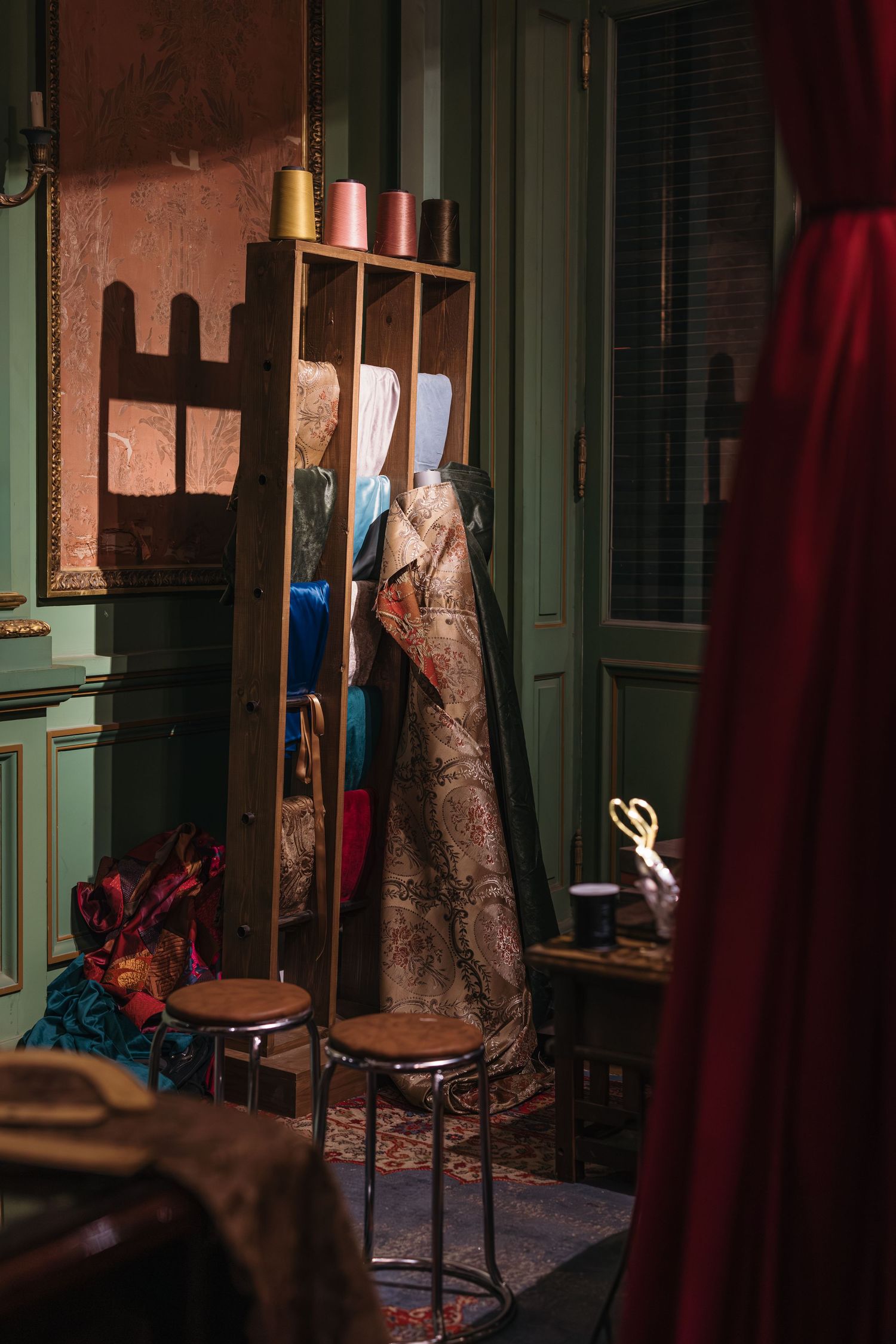 Vintage tailor studio with ornate garments on rack, thread spools, stools, and antique sewing accessories.