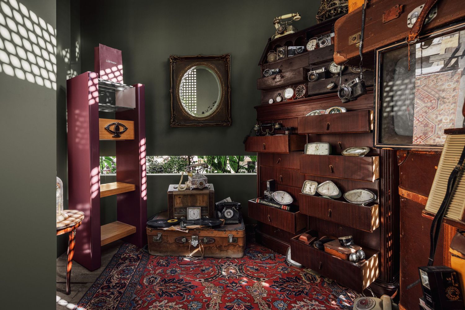 Eclectic vintage room with wooden shelving, antique objects, suitcases, and a rich red patterned rug.